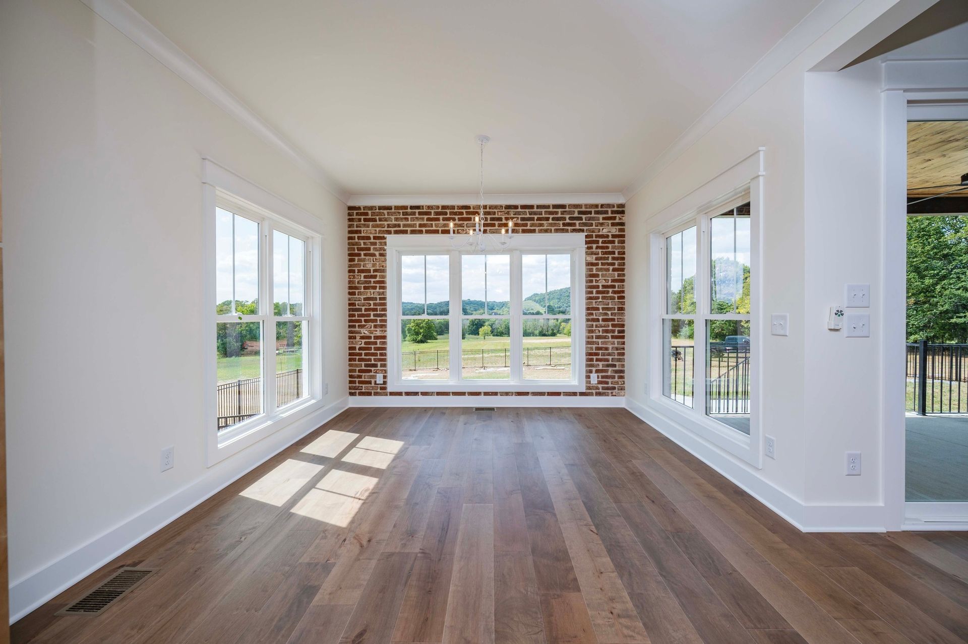 Empty room with hardwood floors, white walls, and brick accent wall, framed by large windows.