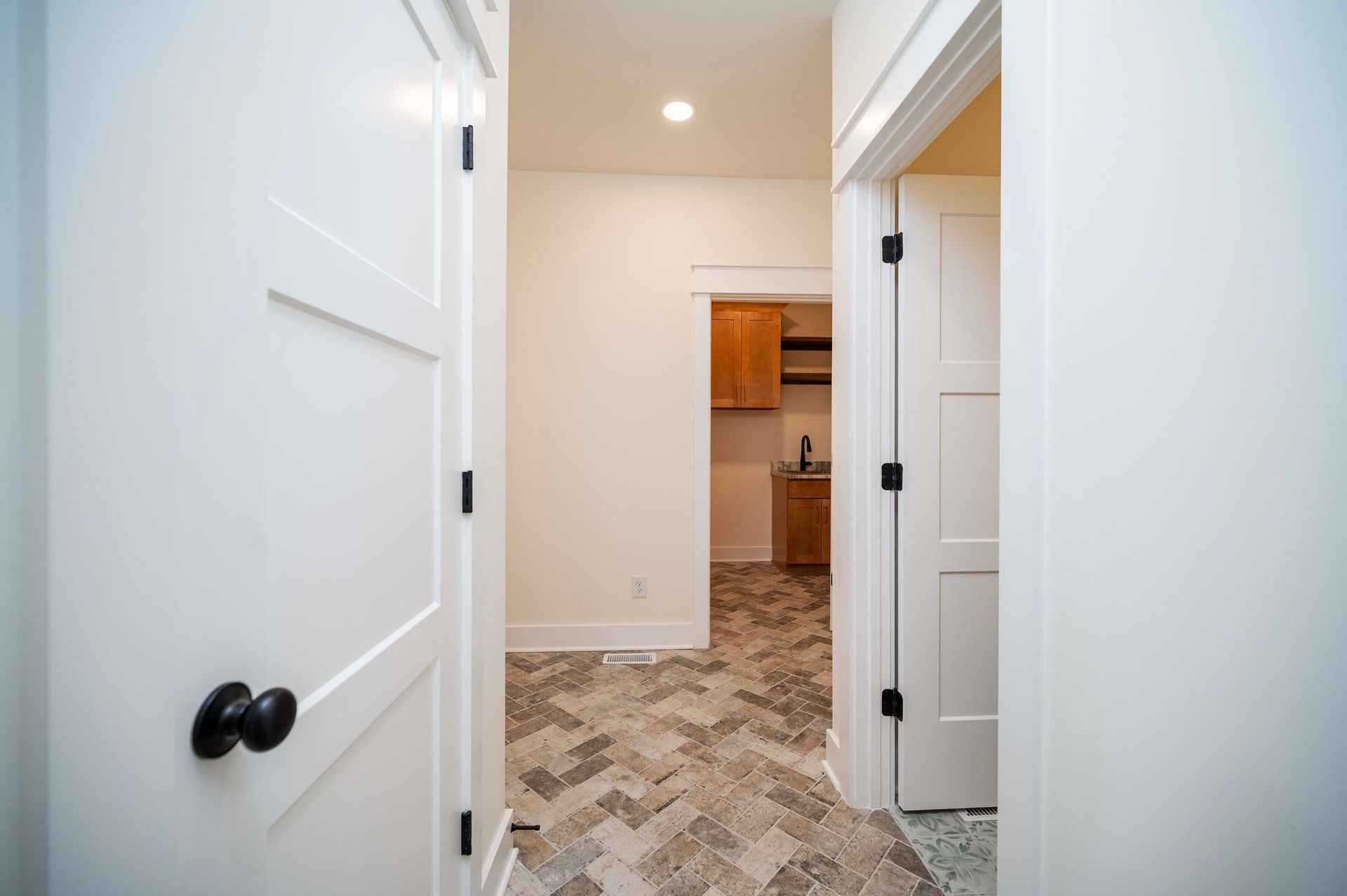White hallway with two doors open to a room with wood cabinets and patterned flooring.