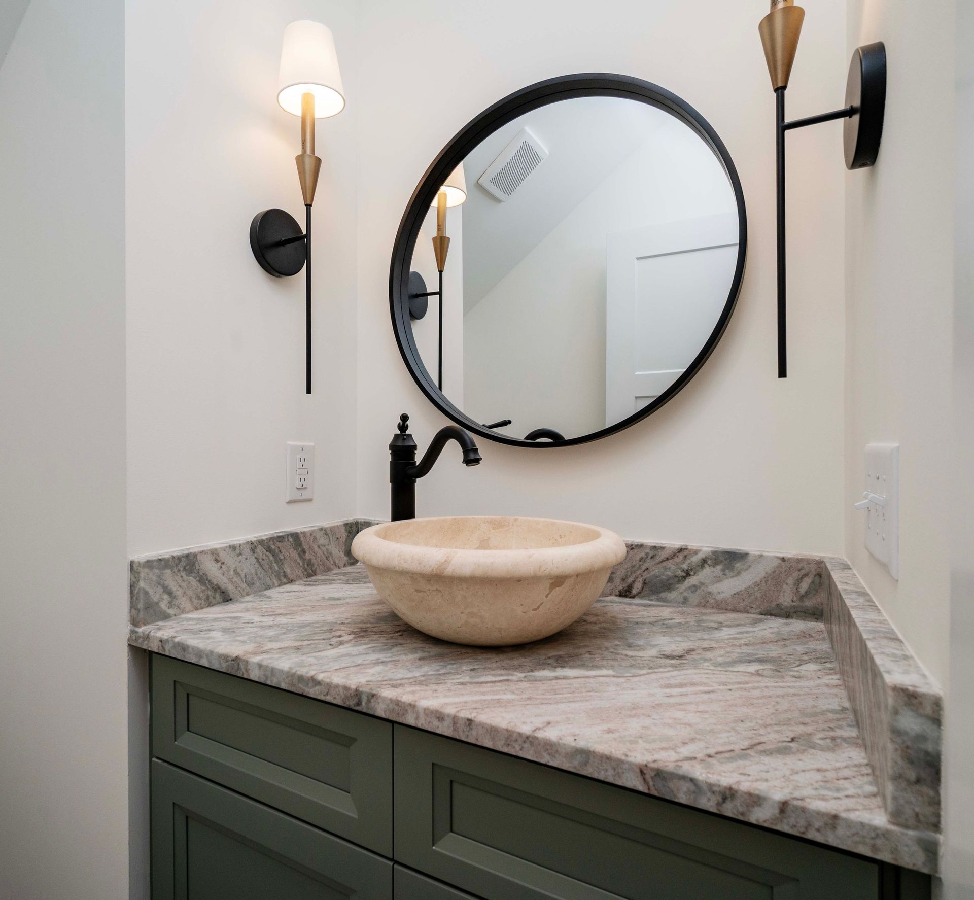 Bathroom with green cabinet, stone countertop, bowl sink, round mirror, and sconces.
