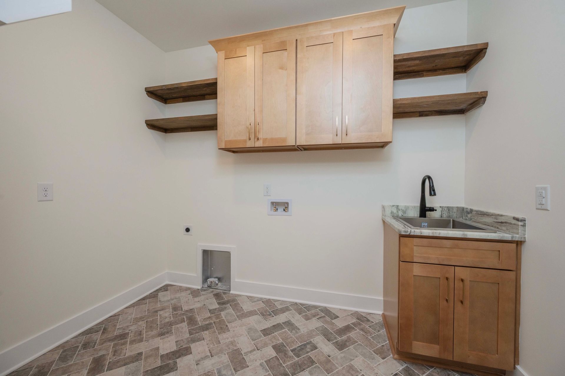 Laundry room with light wood cabinets, floating shelves, sink, and herringbone tile floor.