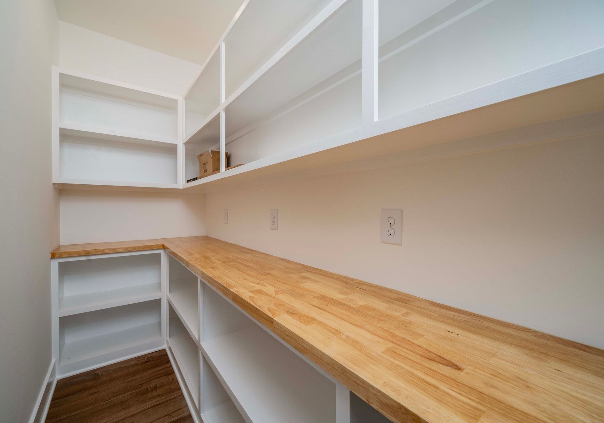 Empty pantry with white shelves, wooden countertop, and an electrical outlet.