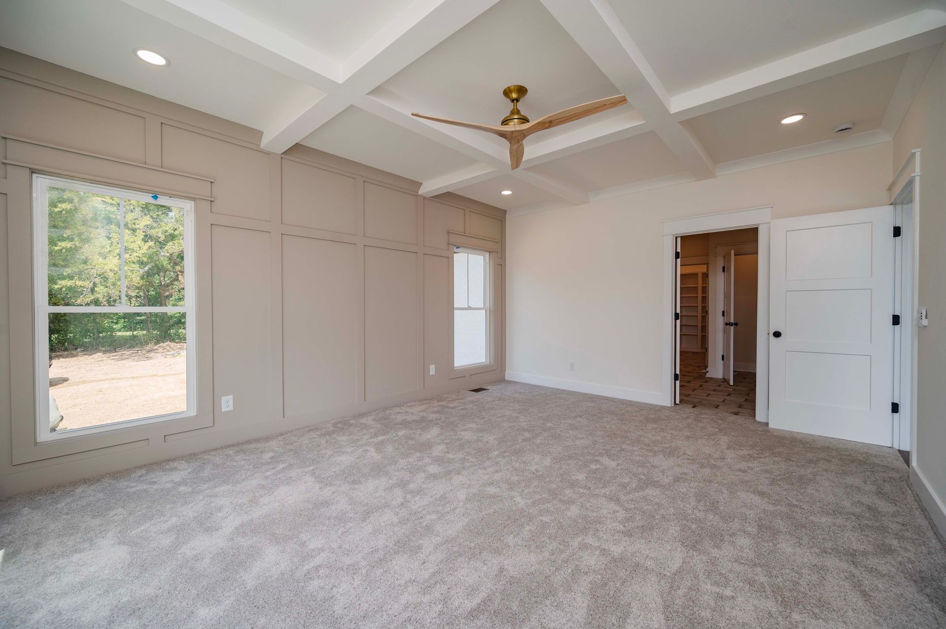 Empty bedroom with light grey walls, beige carpet, and a white coffered ceiling with a gold ceiling fan.