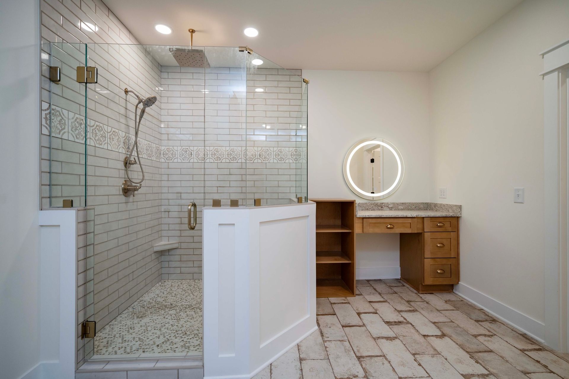 Bathroom with glass shower, vanity with round light, and brick-patterned floor.