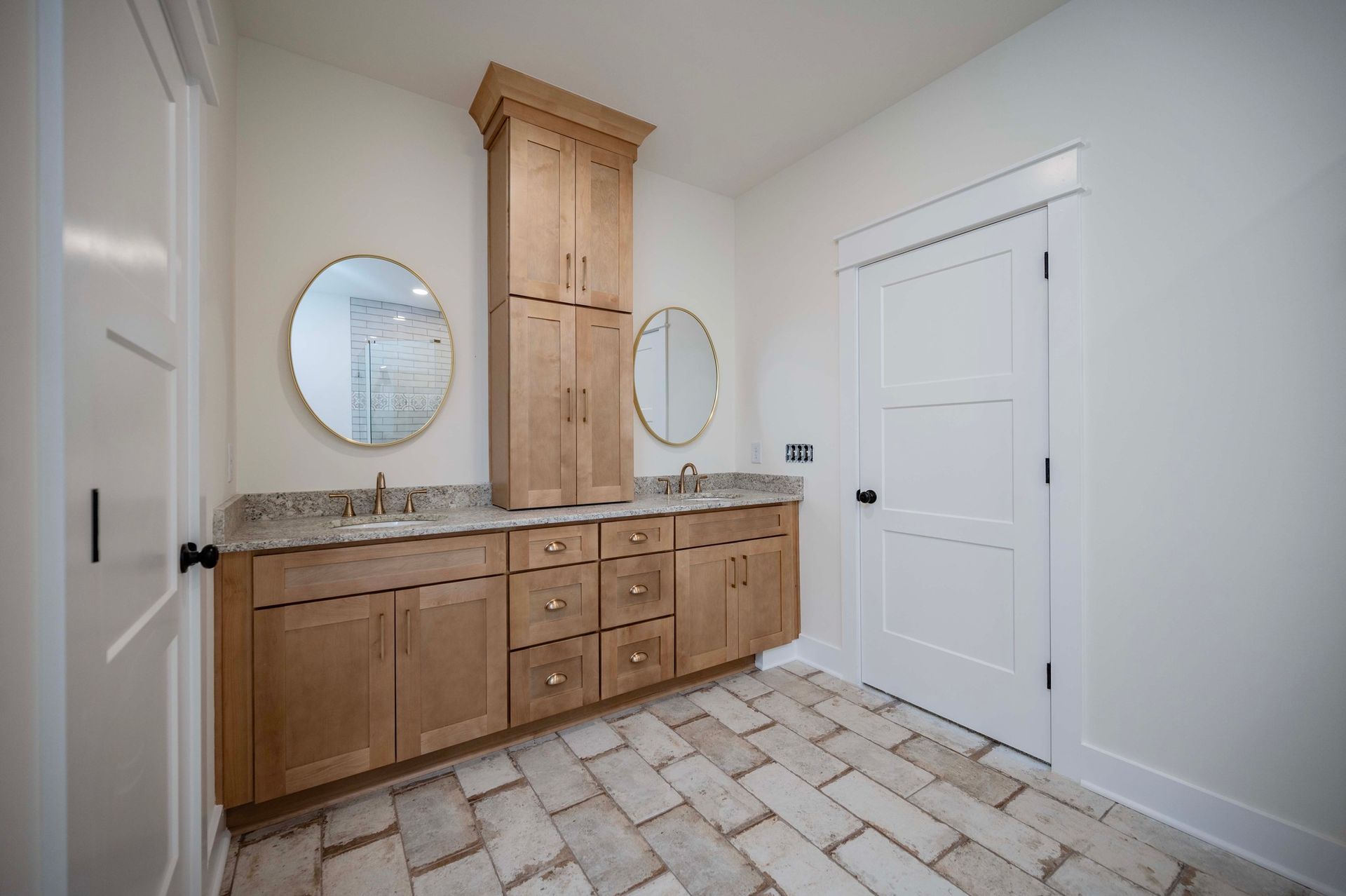 Bathroom with wooden cabinets, beige brick floor, two round mirrors, and white doors.