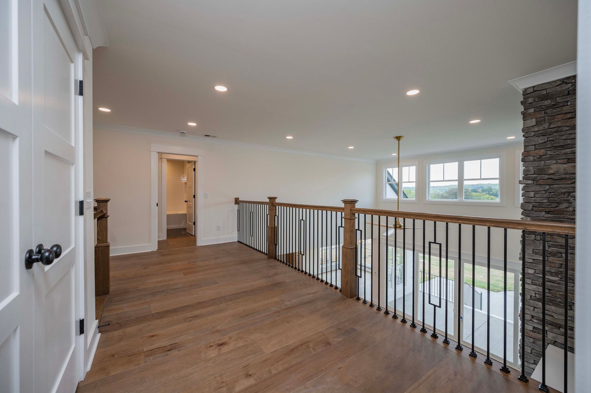 Hallway with hardwood floor and black metal railing, leading to a bathroom and windows.