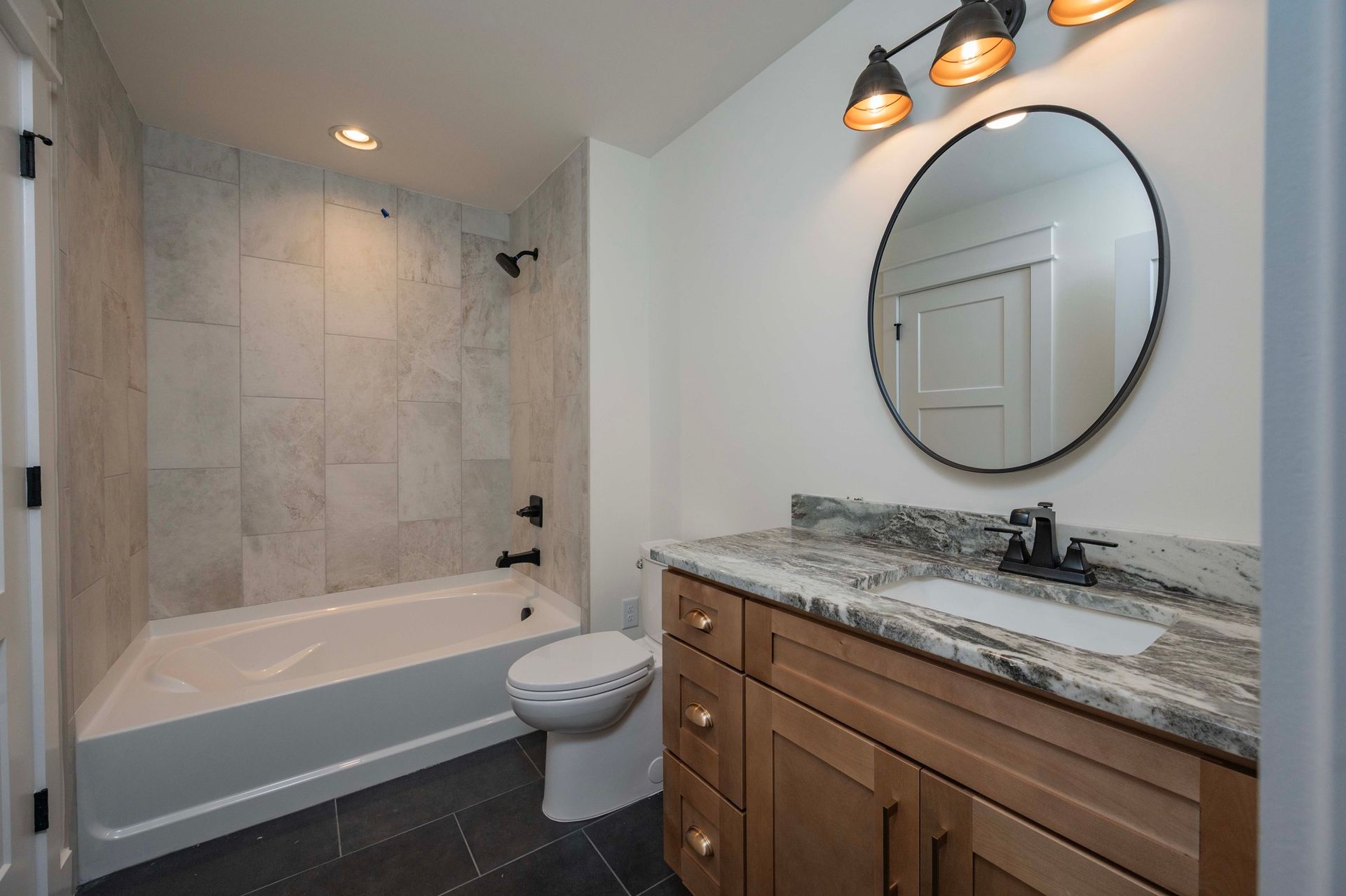Bathroom with white tub, marble tile, brown vanity, black fixtures, and round mirror.
