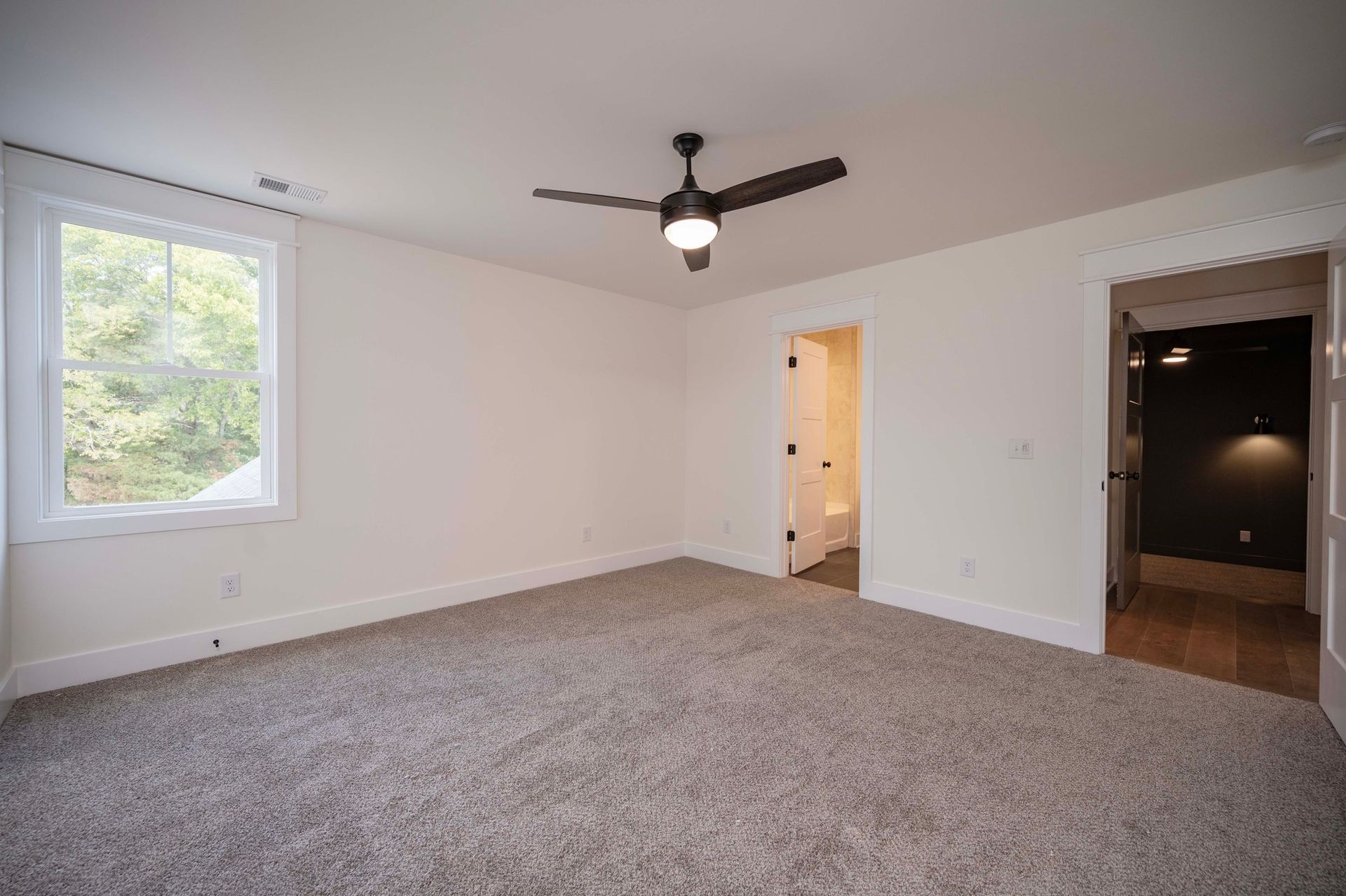 Empty bedroom with light carpet, white walls, ceiling fan, and doorway to a bathroom.