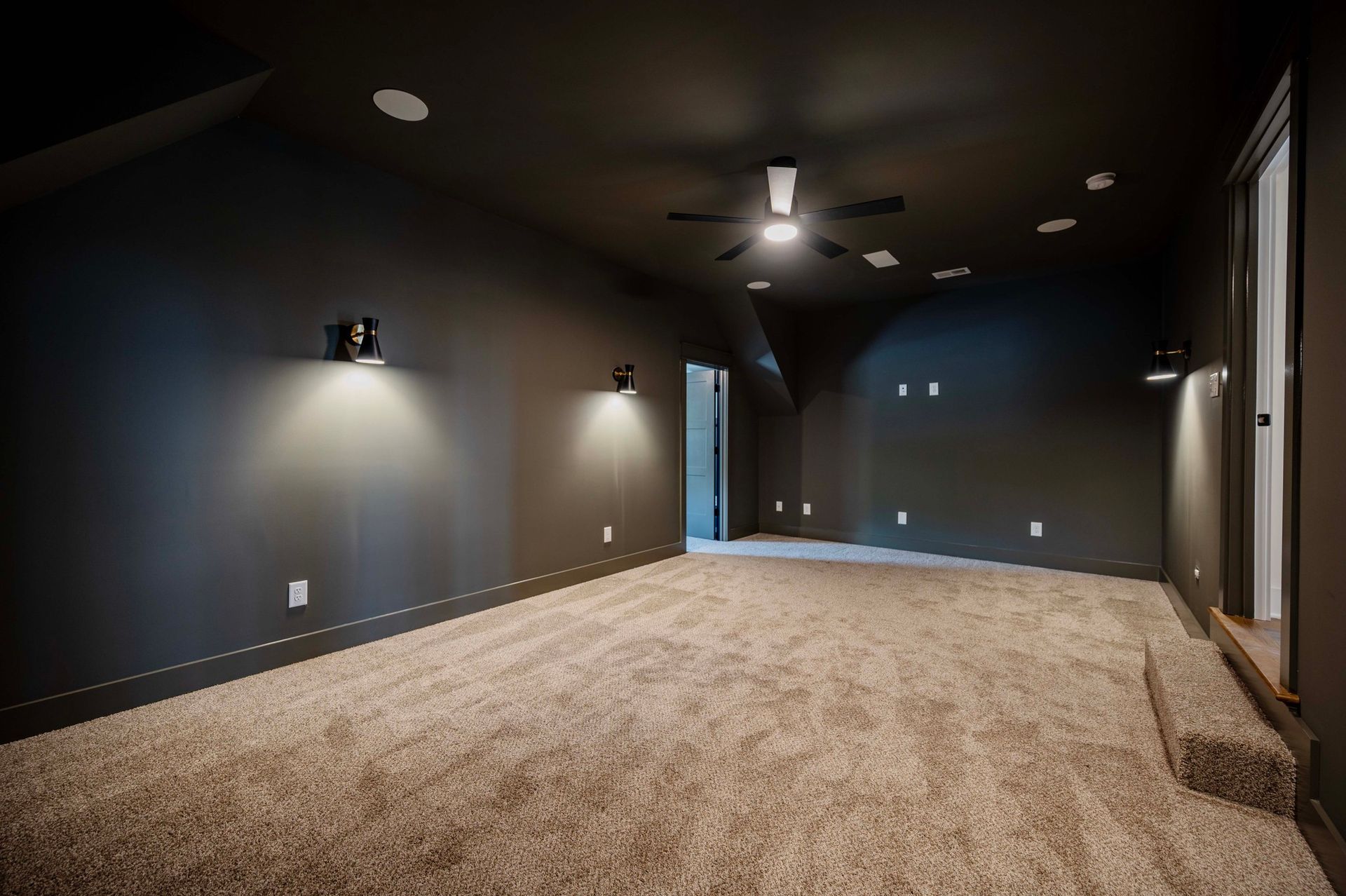 Empty dark-walled room with beige carpet, recessed lighting, wall sconces, ceiling fan, and open doorways.
