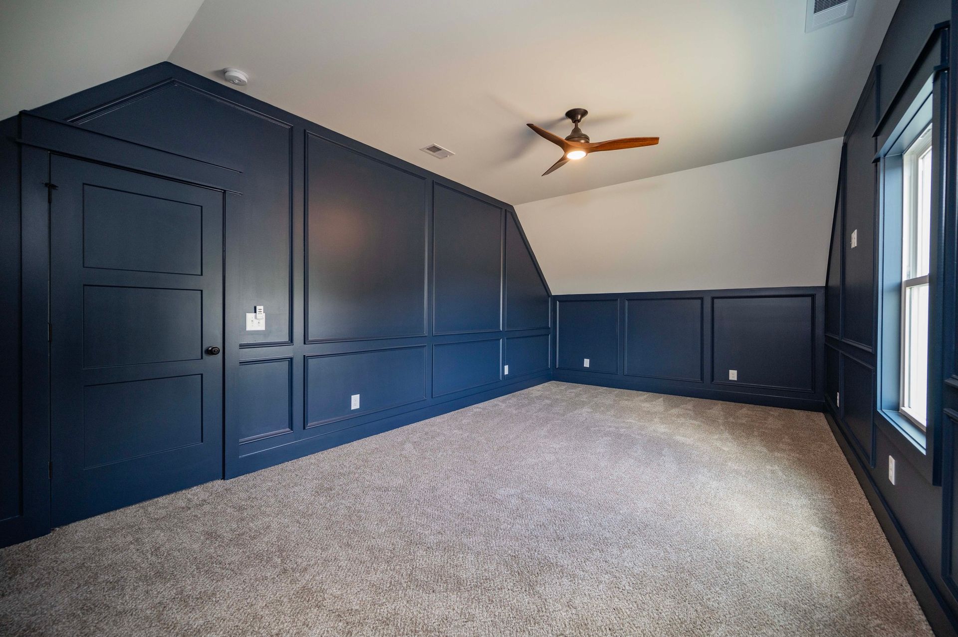 Empty room with blue paneled walls, white ceiling, and beige carpet. A ceiling fan and window are visible.
