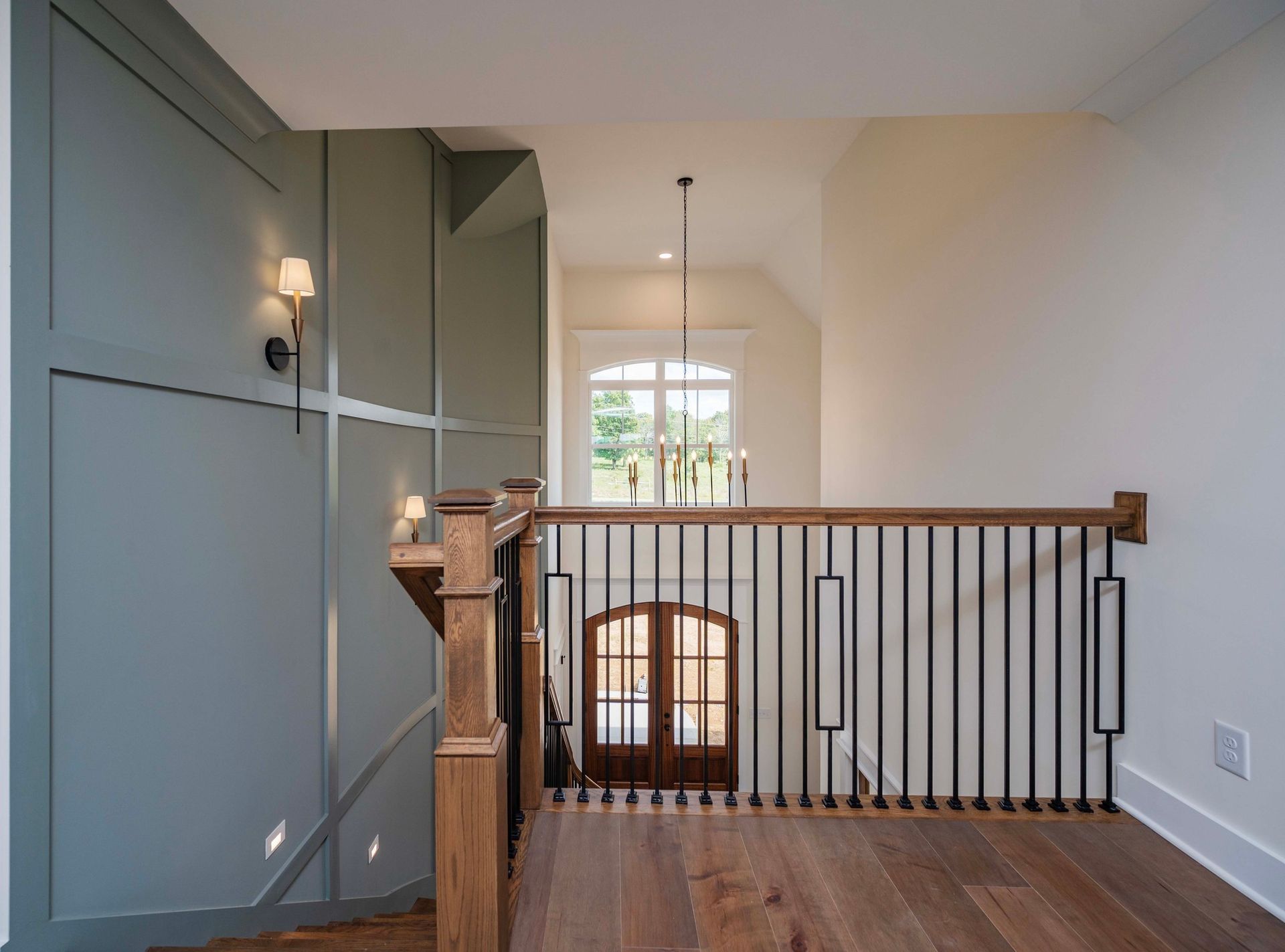 Interior view of a landing with stairs, wood floors, and a railing. Green accent wall with sconces, and a window at the end.