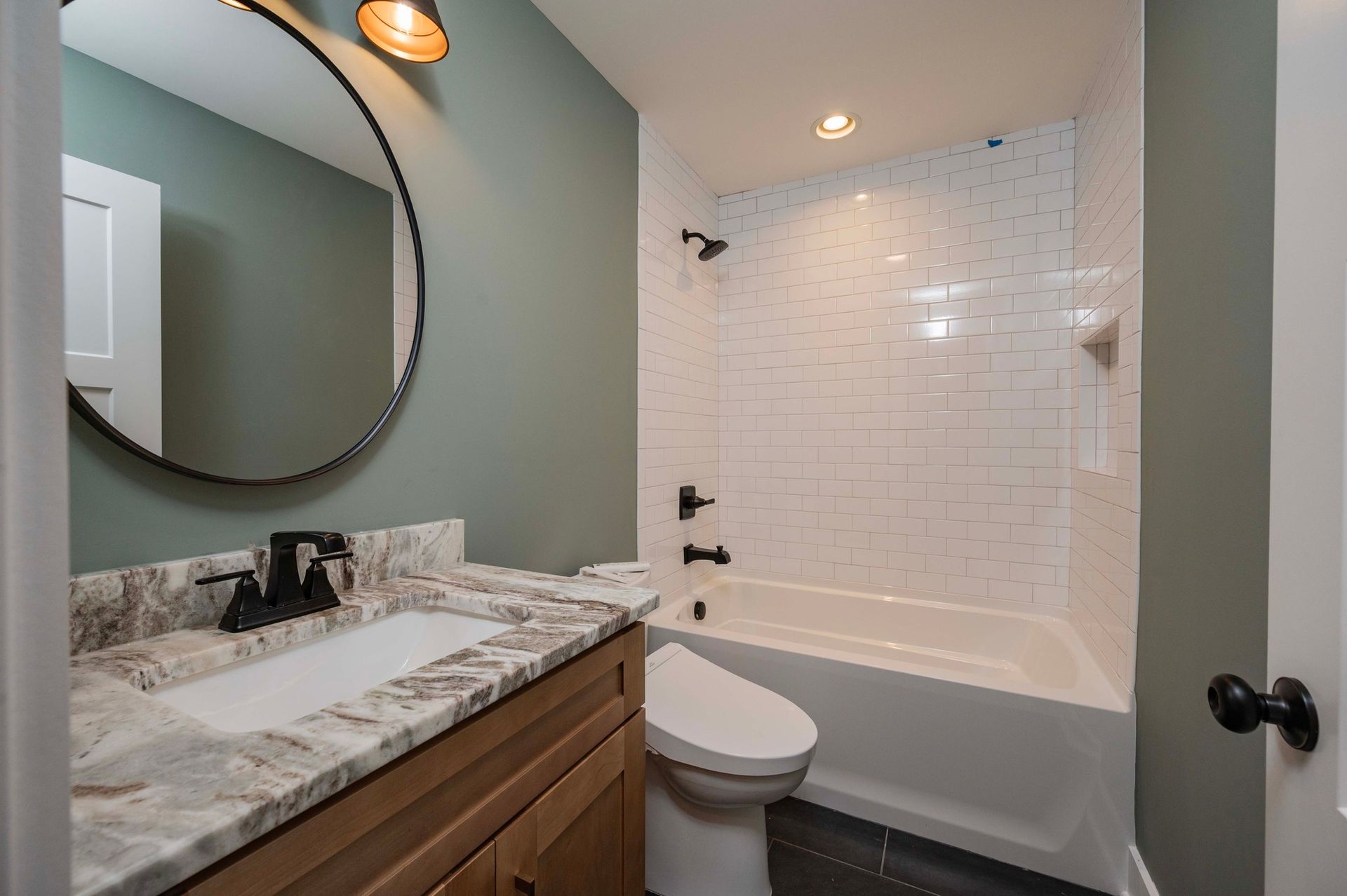 Bathroom with a white bathtub, toilet, and sink. Walls are green with a round mirror.