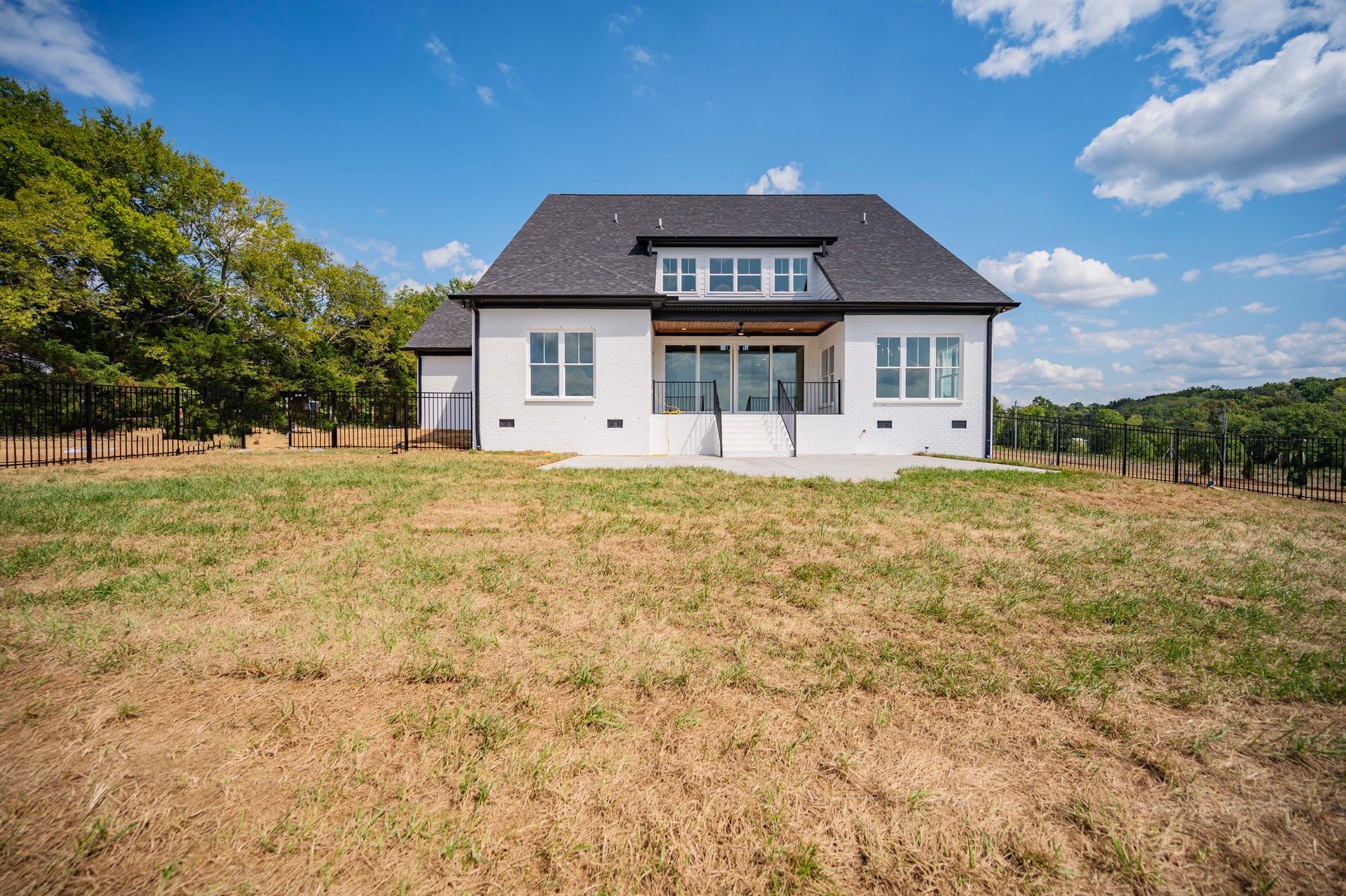 White house with black roof, large windows, and a patio, set in a grassy field under a blue sky.