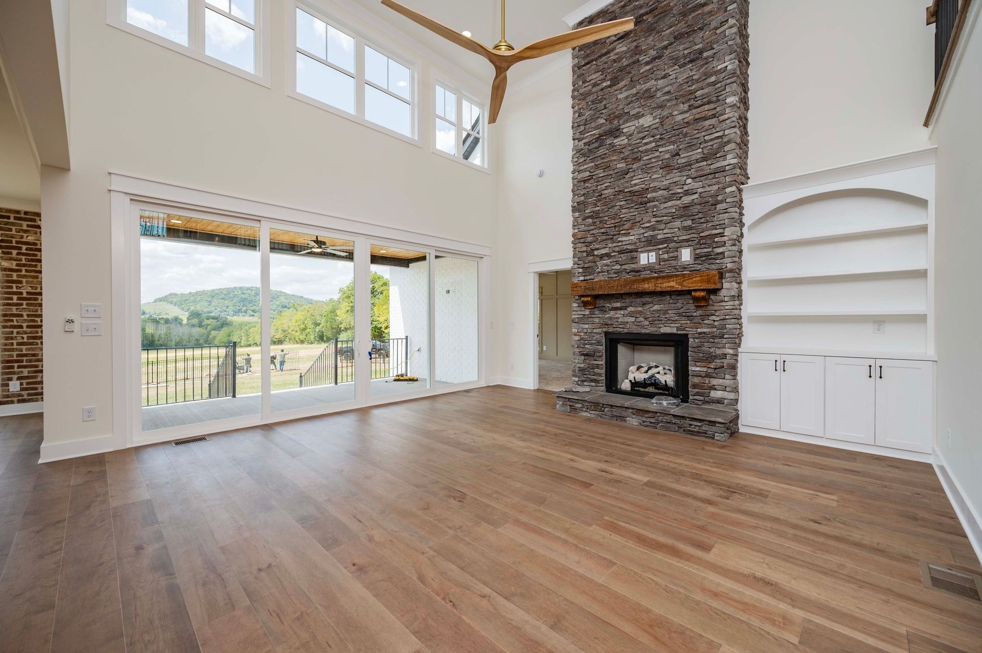 Spacious living room with hardwood floors, a stone fireplace, and large windows overlooking a green landscape.