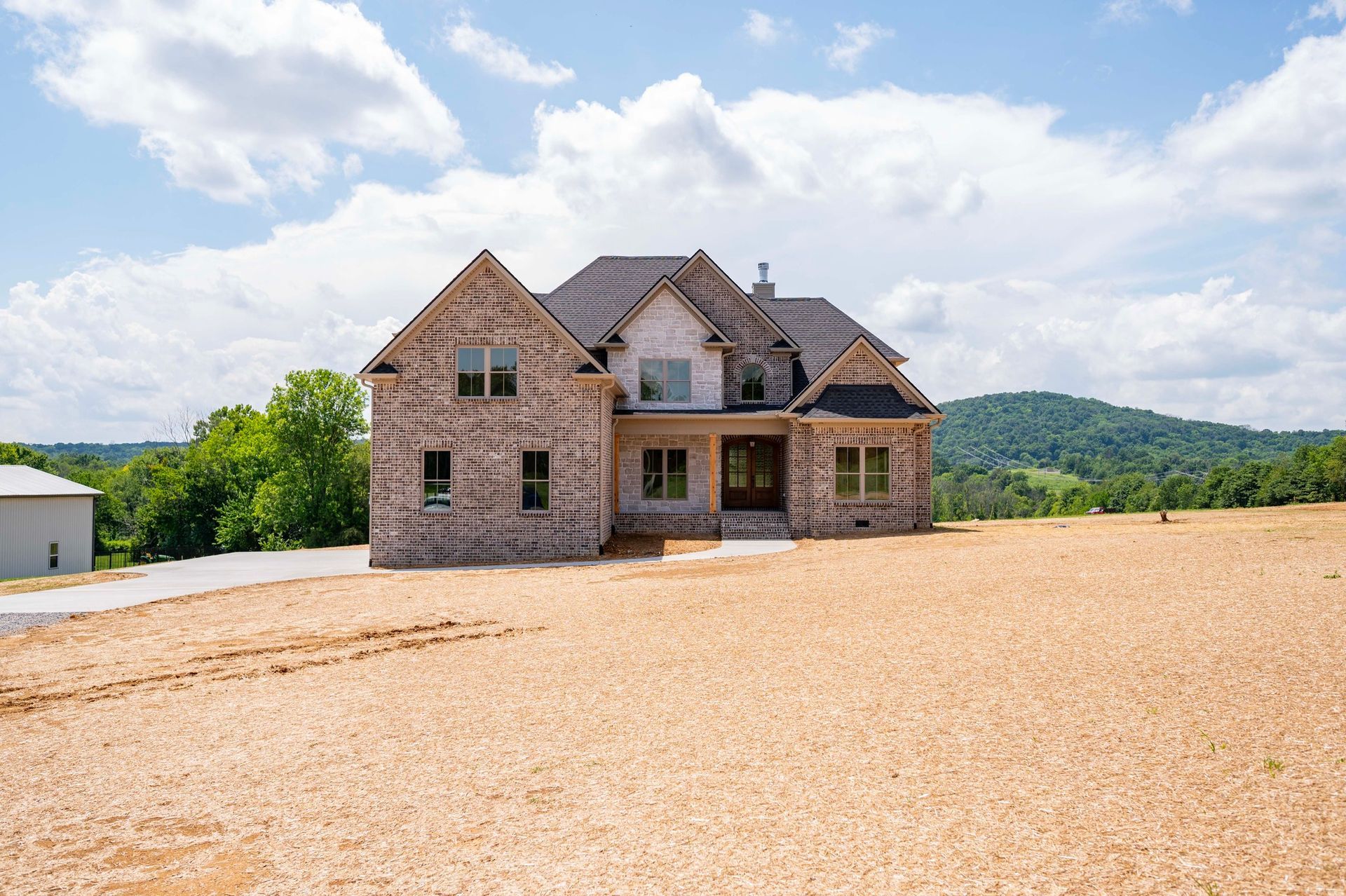 New two-story brick house with brown roof on a large cleared lot, hills in background.