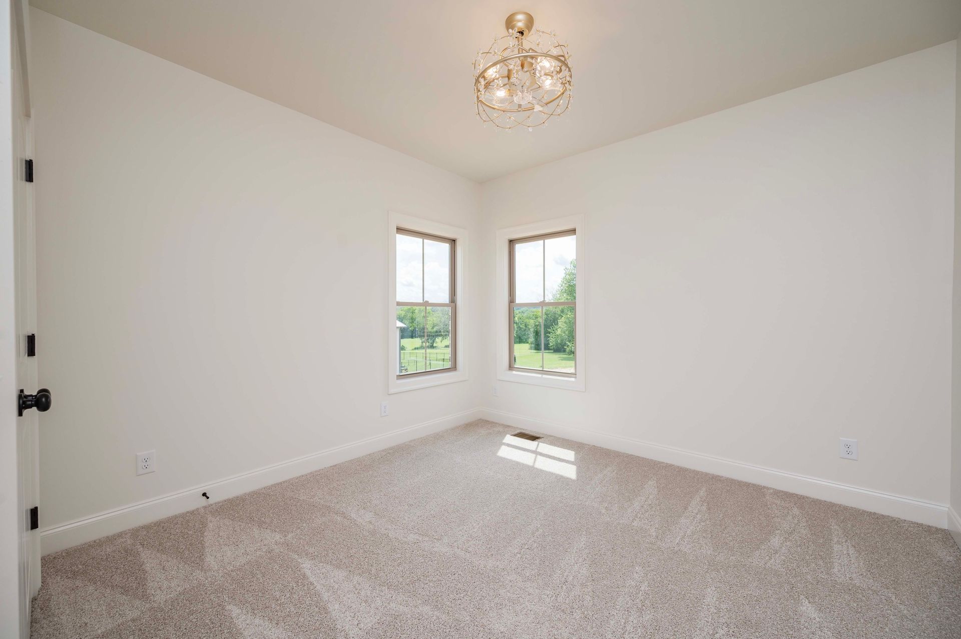 Empty room with off-white walls, carpet, two windows, and a chandelier; door visible.