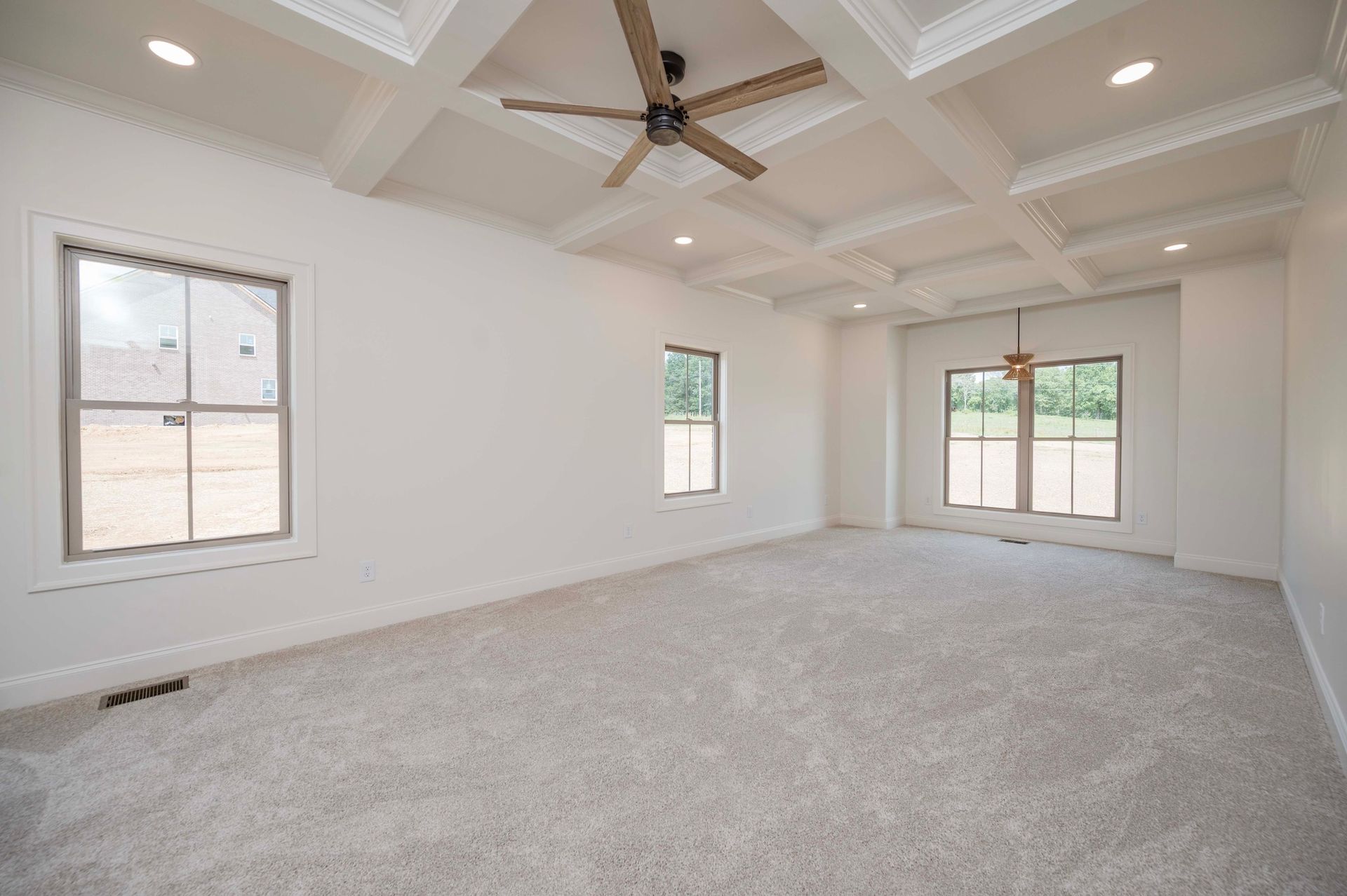 Empty, bright bedroom with gray carpet, white walls, and a coffered ceiling. Windows provide natural light.