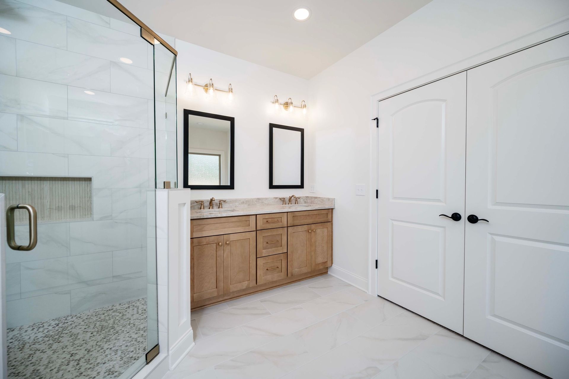 A modern, bright bathroom with marble tile and a glass shower. Light wood vanity with two mirrors.