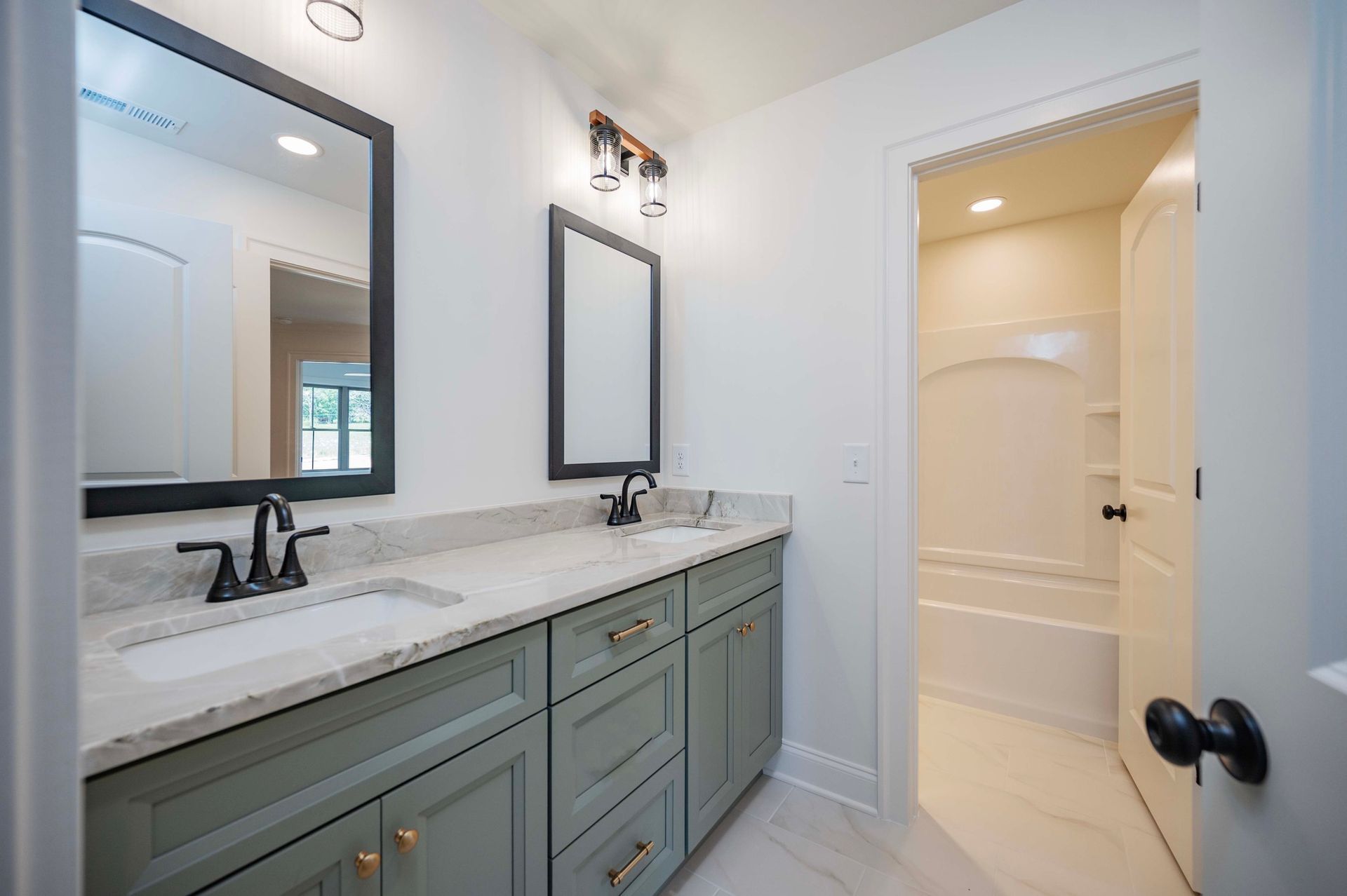 Bathroom with sage green vanity, gray countertop, black fixtures, and white tiled floor. Doorway to a bathtub.