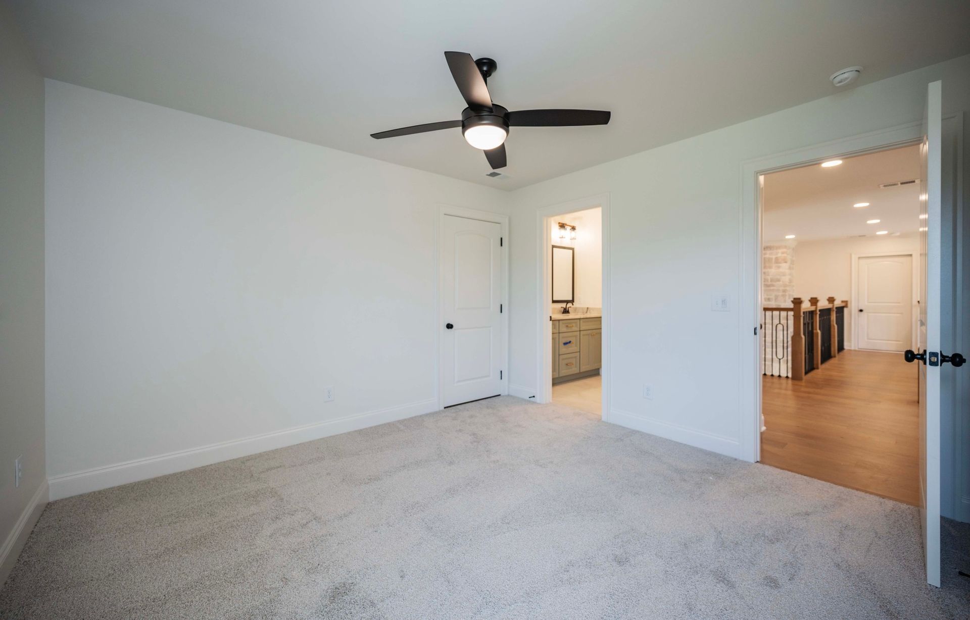 Empty room with light gray carpet, white walls, black ceiling fan, and doors leading to a bathroom and hallway.