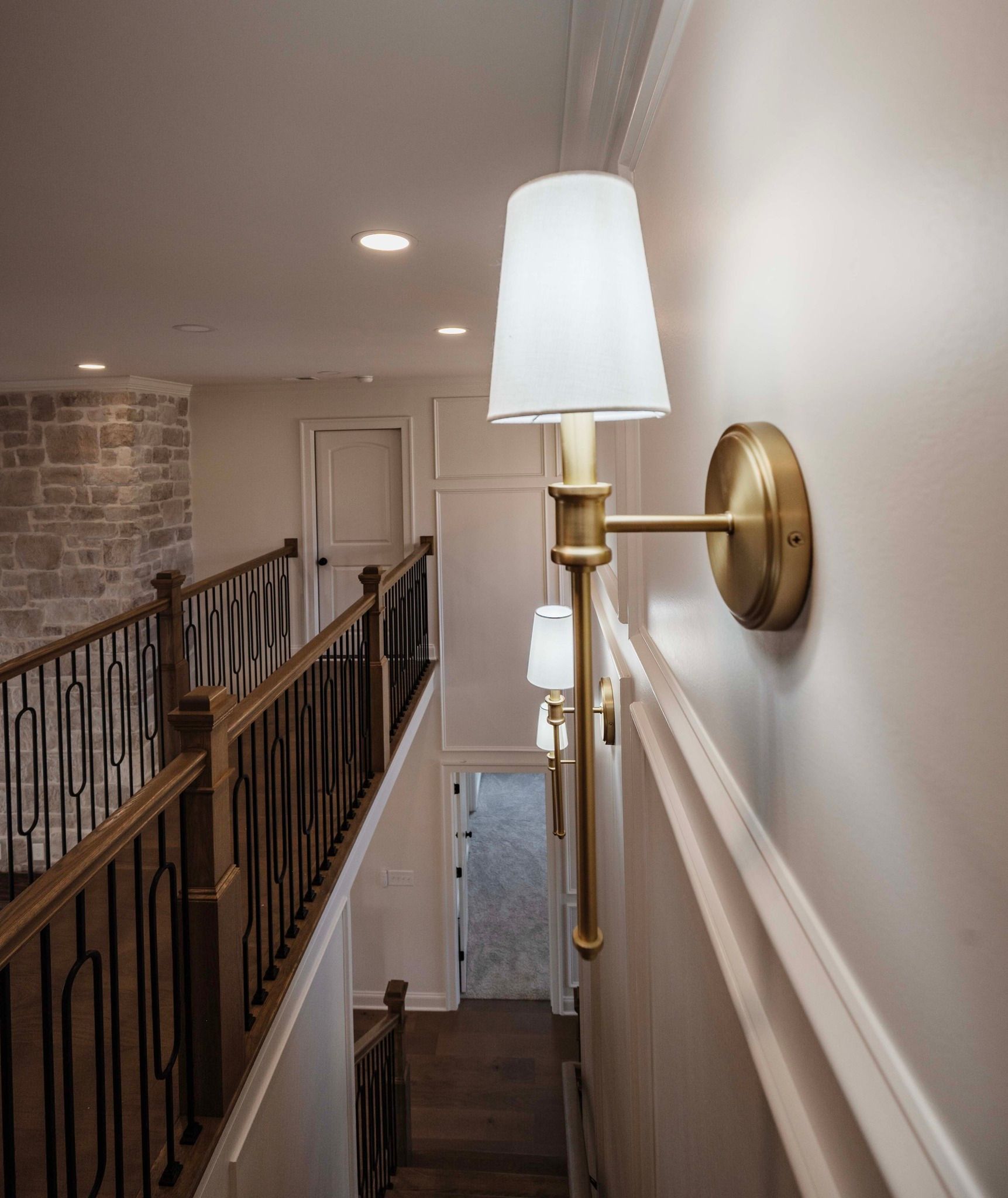 Staircase with gold sconces on the wall. Wooden railing with black details. Light-colored walls and flooring.