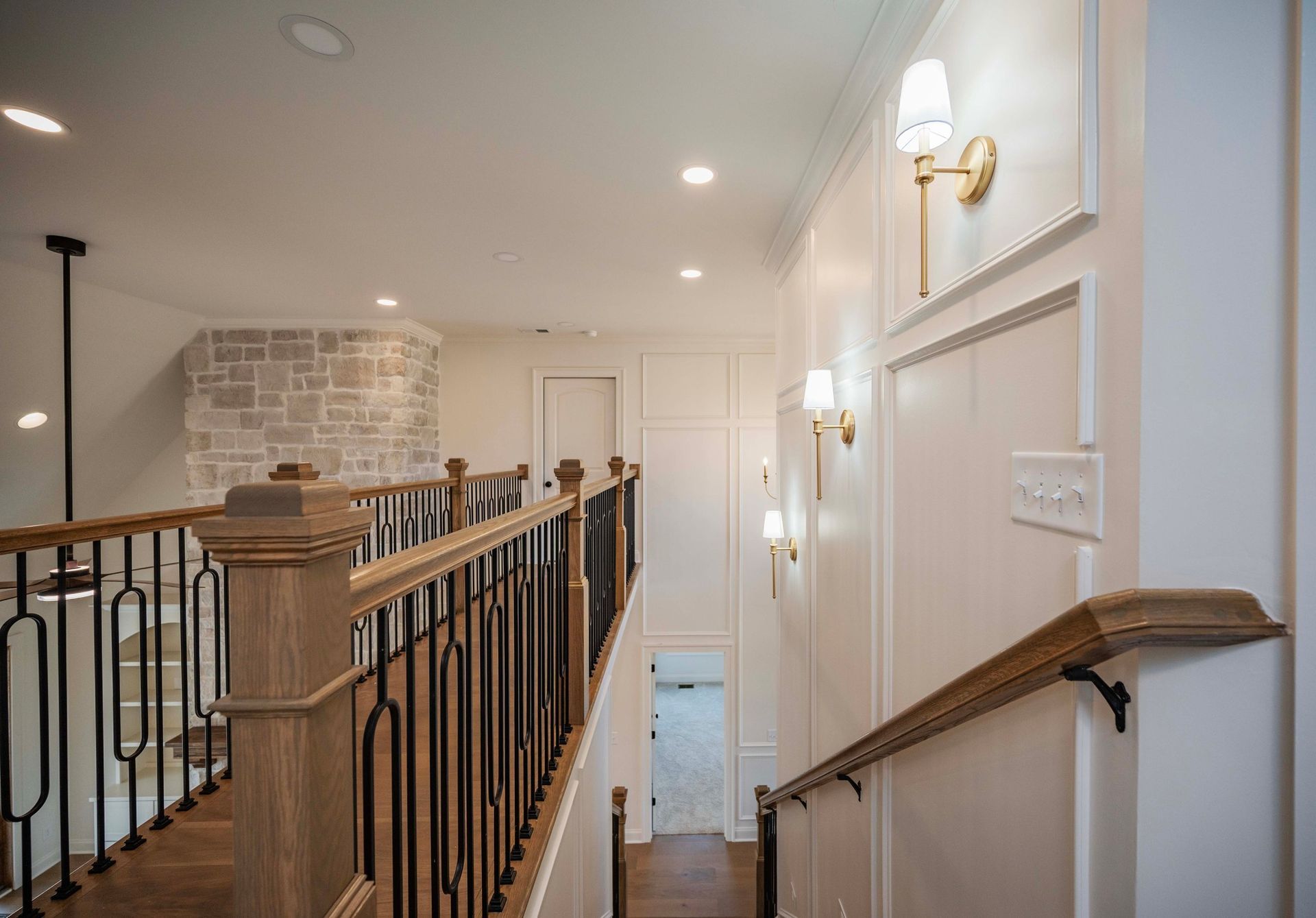 Interior view of a hallway with a staircase, featuring white walls, sconces, and a wooden railing.