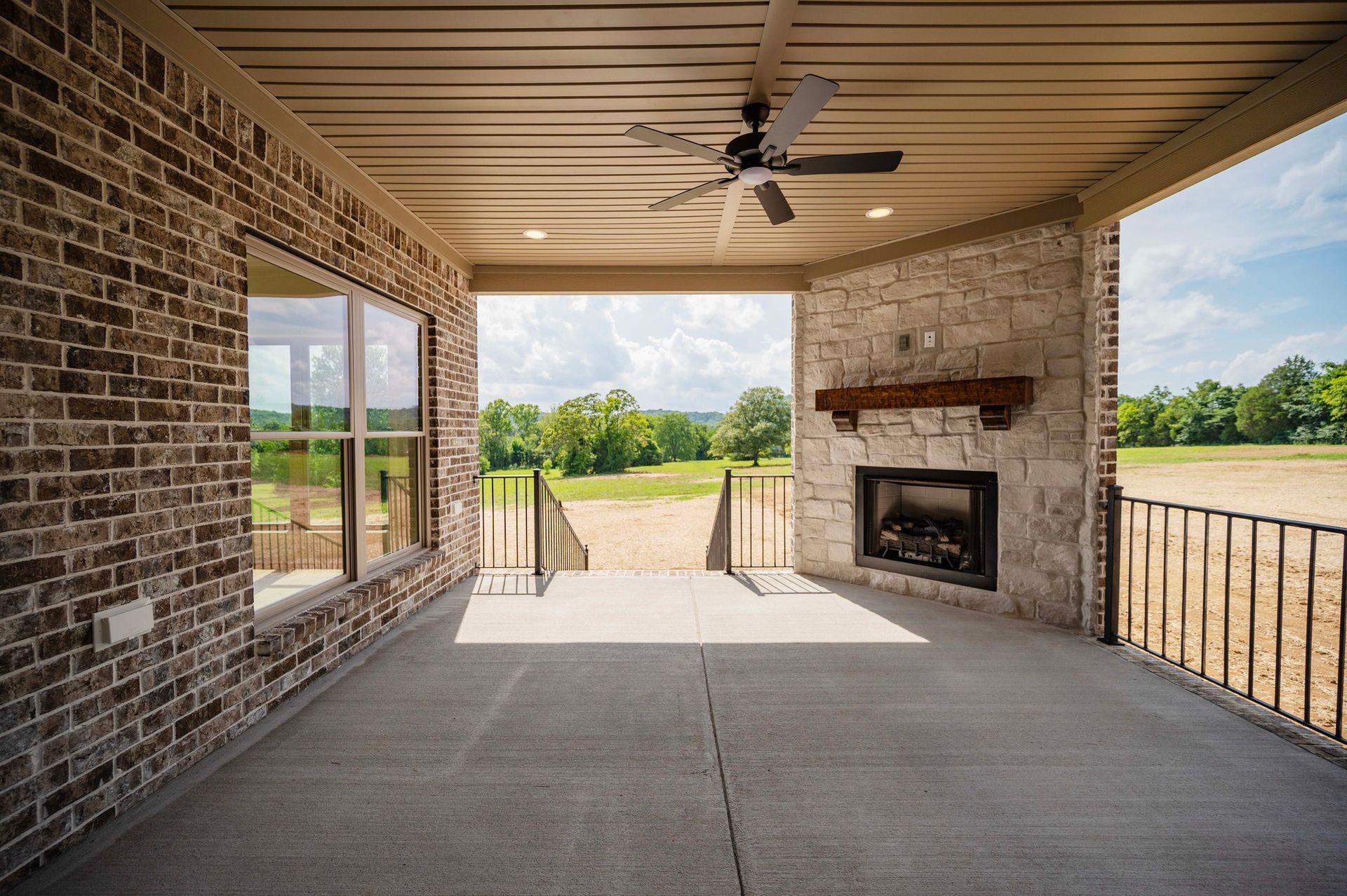 Covered porch with fireplace, brick and stone walls, ceiling fan, and view of a green field.