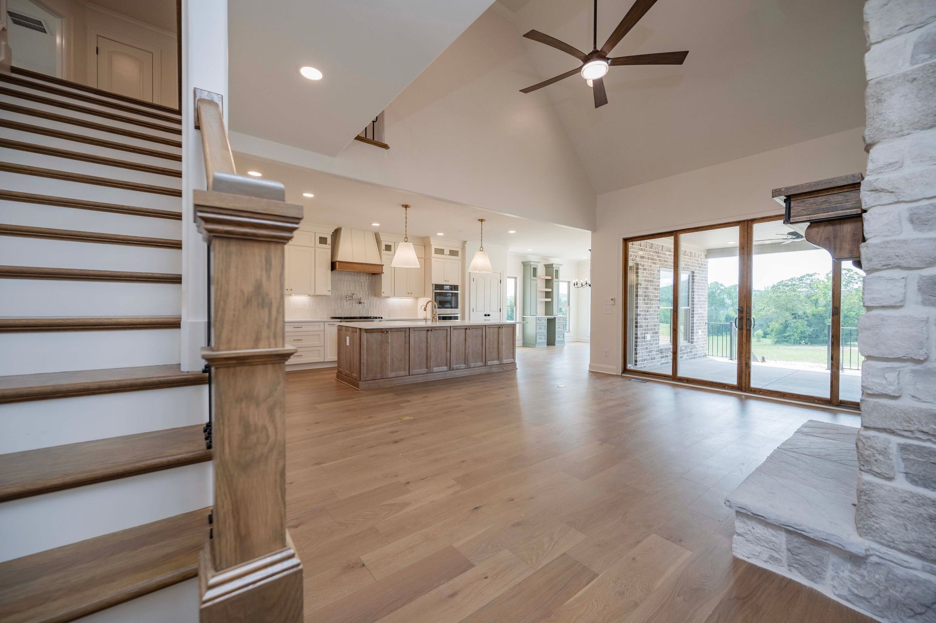 Interior of a spacious, light-filled living area with hardwood floors, kitchen, and stone fireplace; stairs on the left.