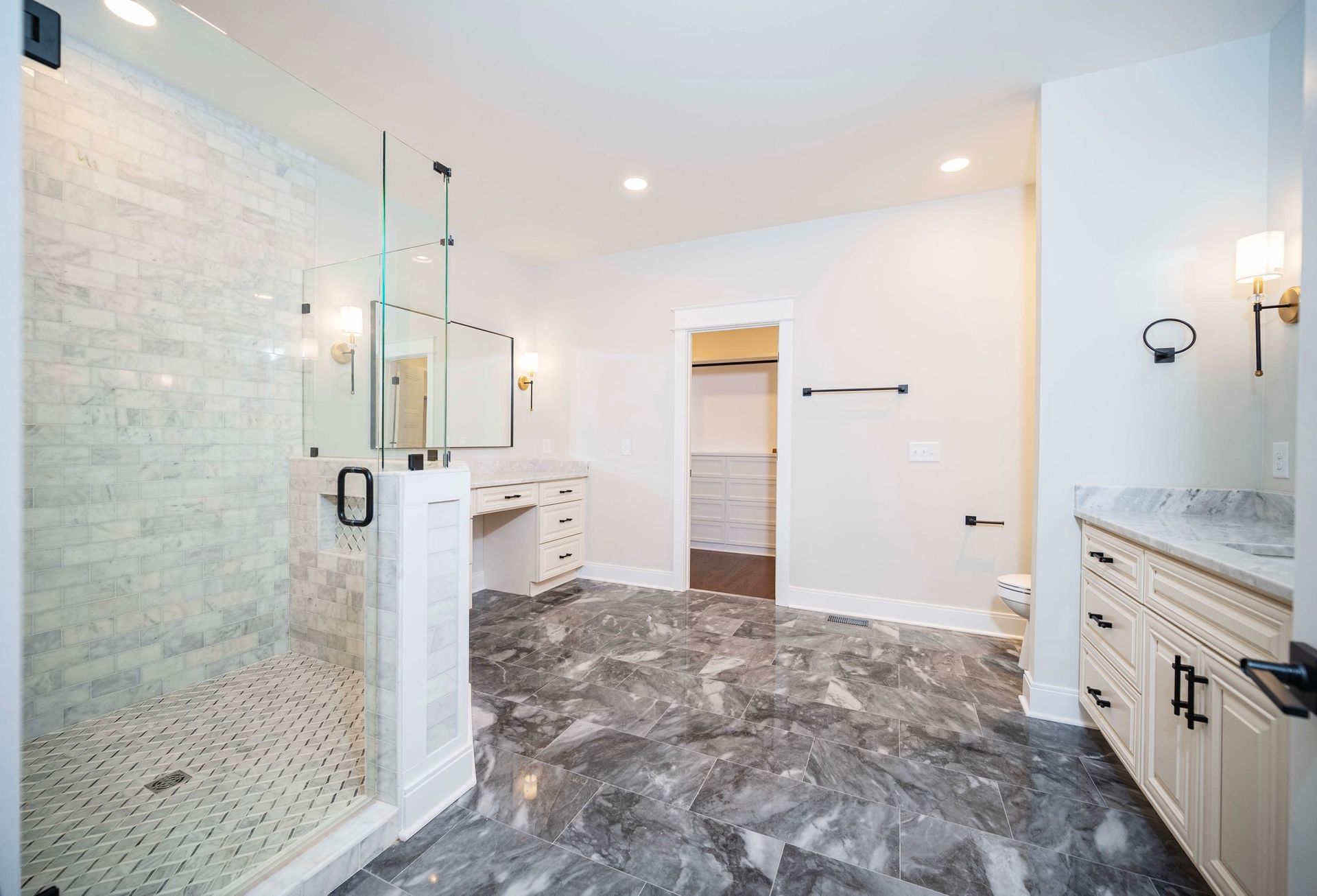 Spacious bathroom with glass shower, vanity, white cabinets, and dark gray marble flooring.