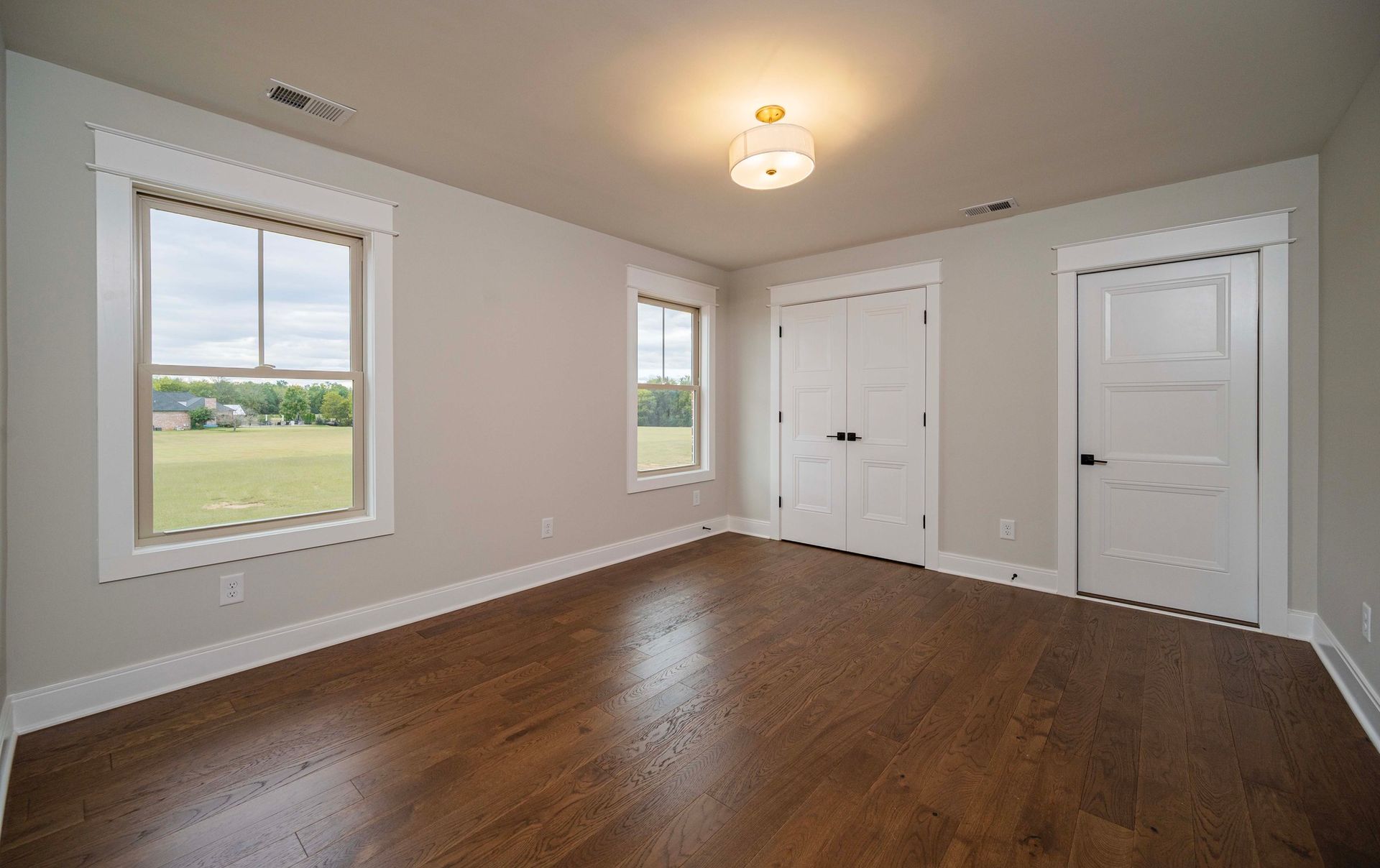 Empty bedroom with wood floors, two windows, two doors, and a light fixture.