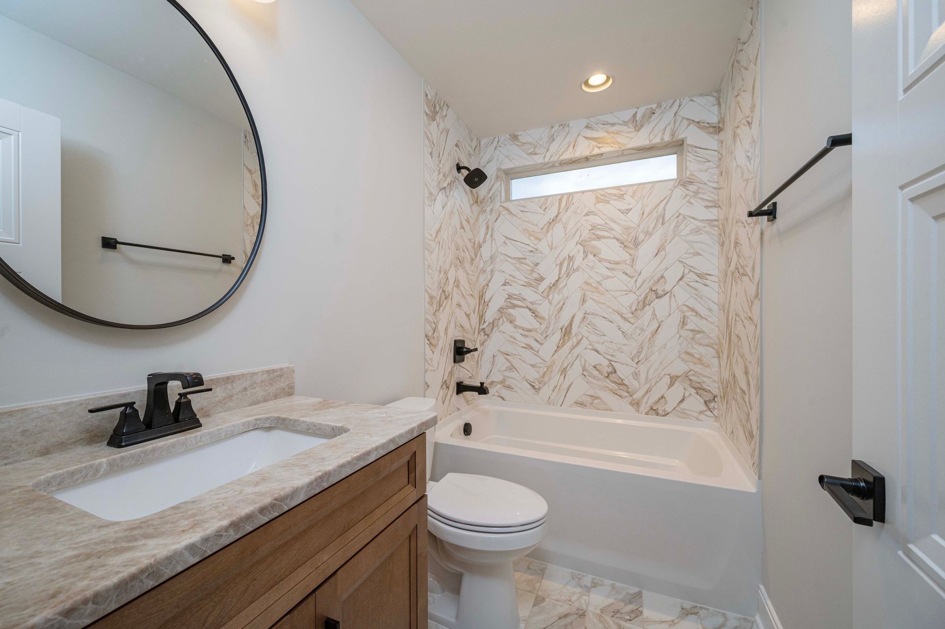 Bathroom with a white tub, toilet, and vanity, and a patterned tile accent wall with a small window.