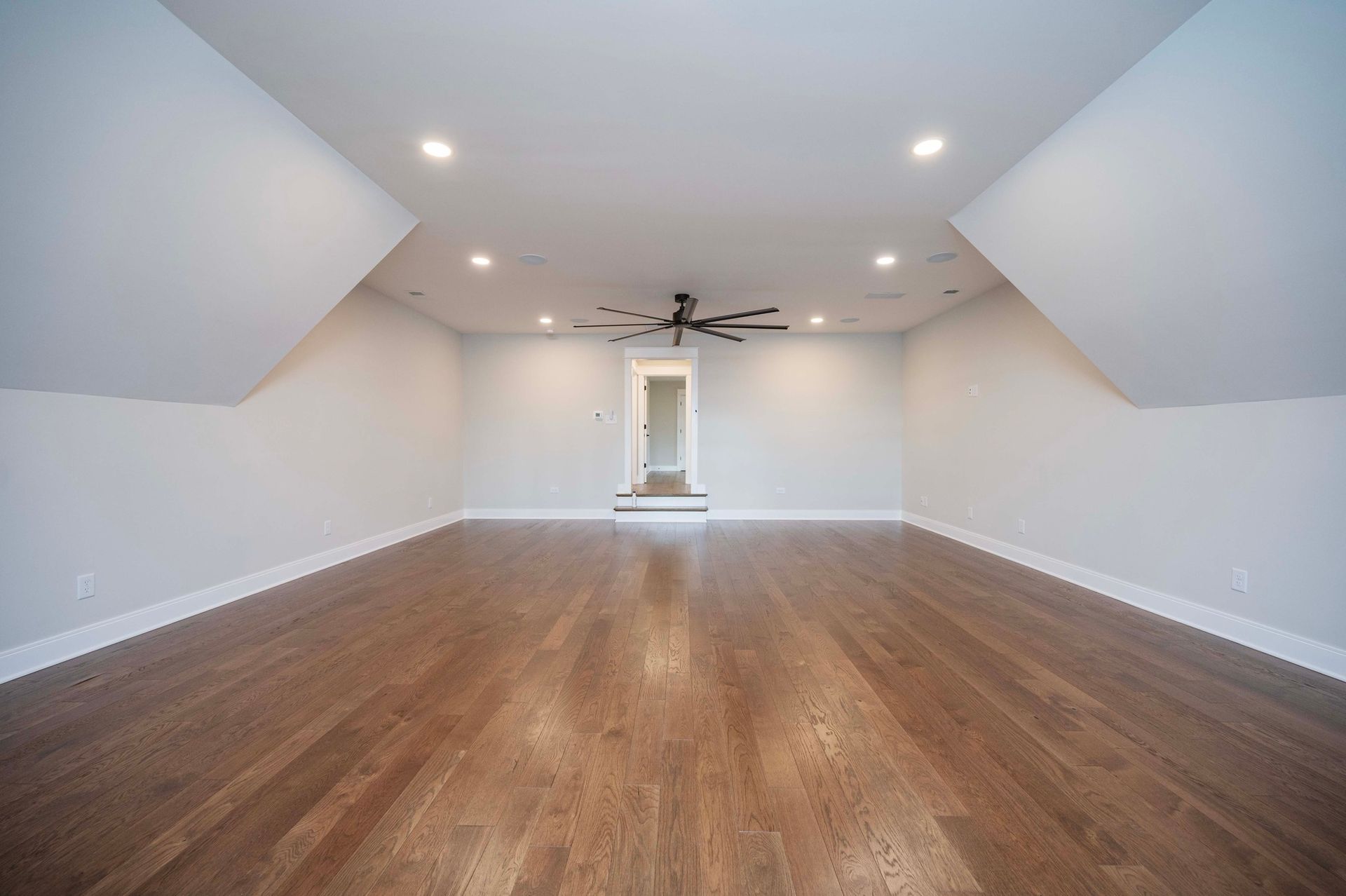 Empty room with hardwood floors, white walls, angled ceiling, and recessed lighting. A doorway is visible.