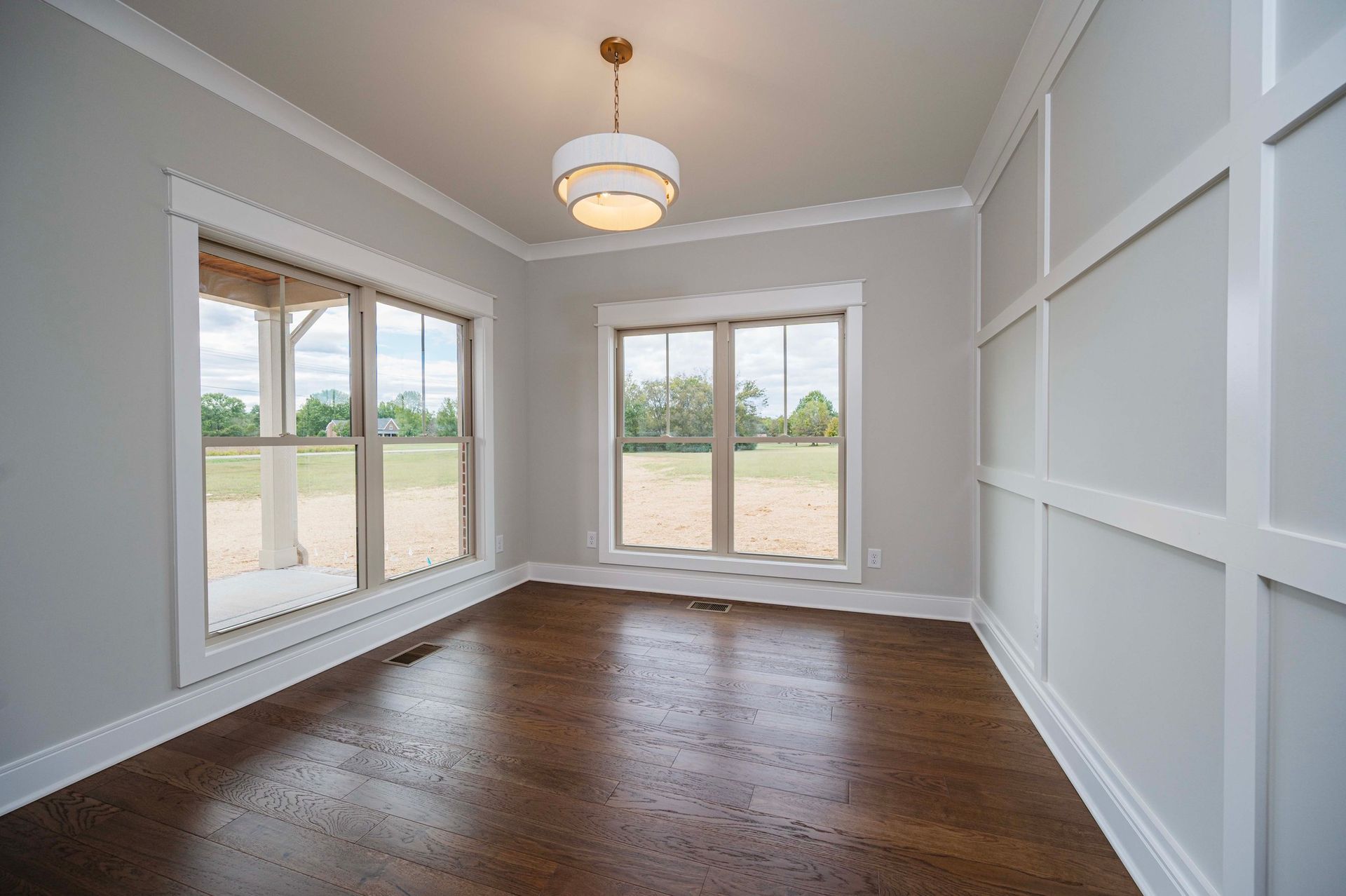 Empty room with hardwood floor, two windows, wainscoting, and a hanging light fixture.