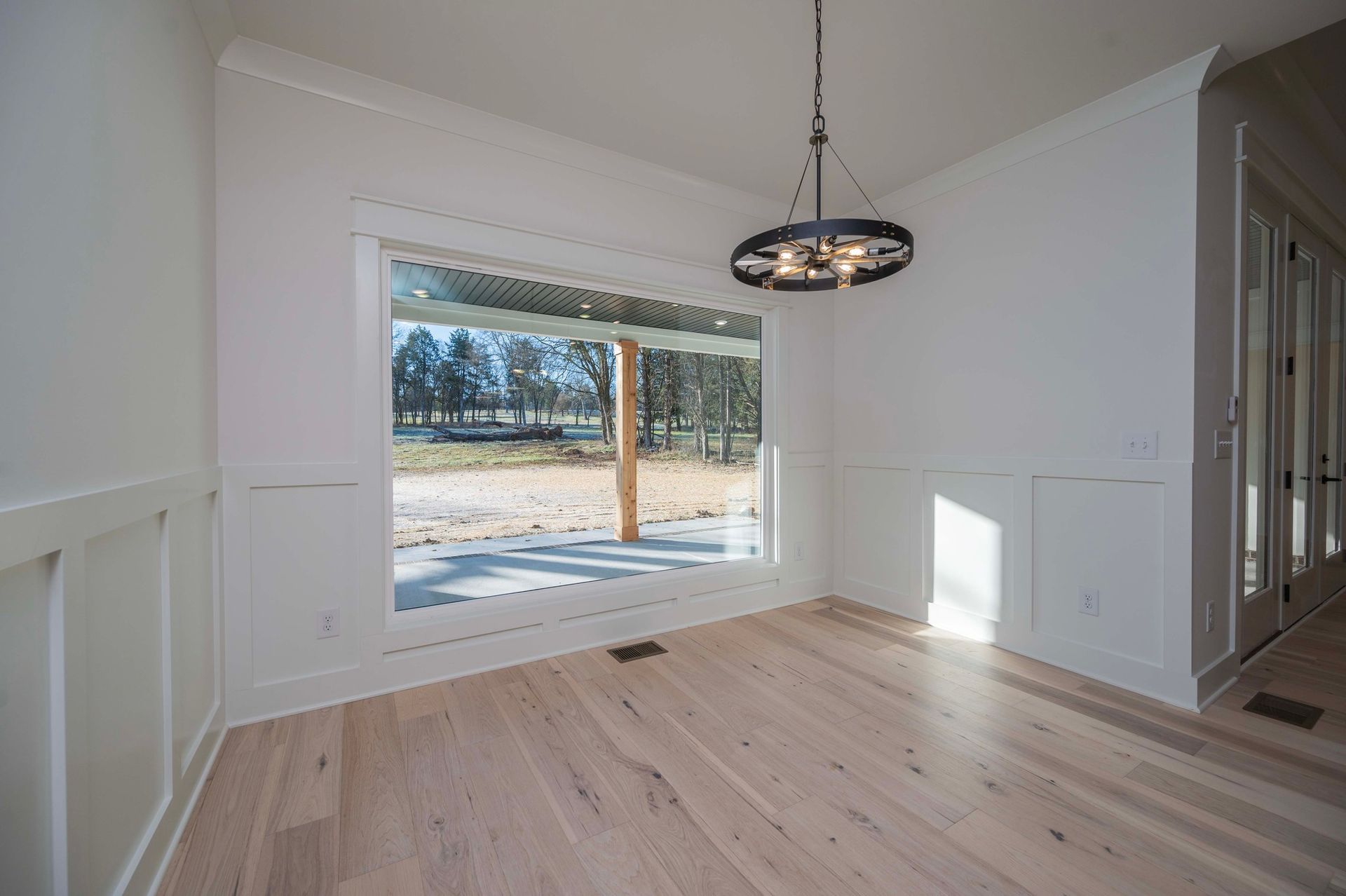 Dining room with hardwood floors, large window, and chandelier. White walls with paneling.