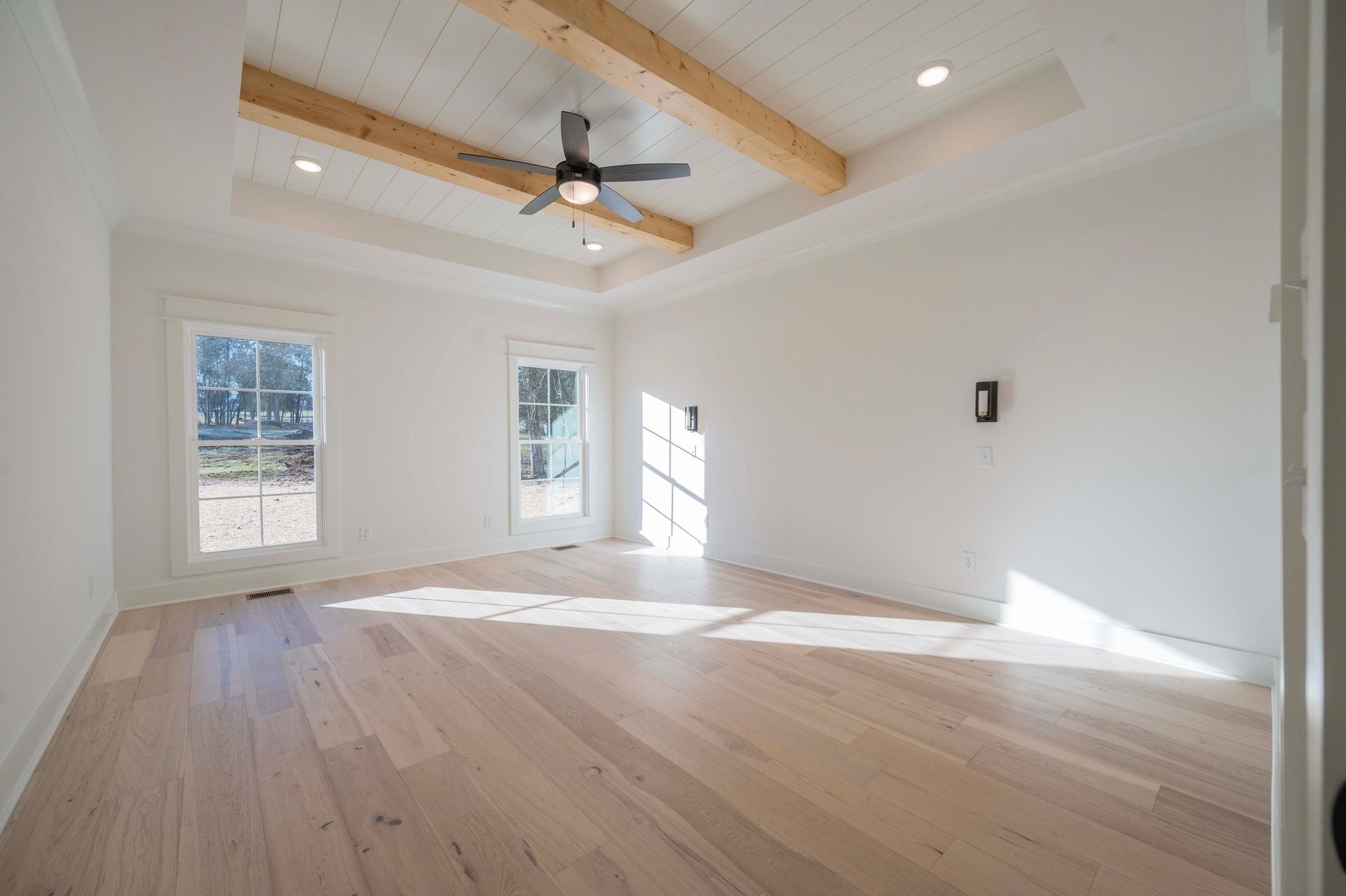 Empty room with light wood floors, white walls, and a recessed ceiling with wood beams and a ceiling fan.