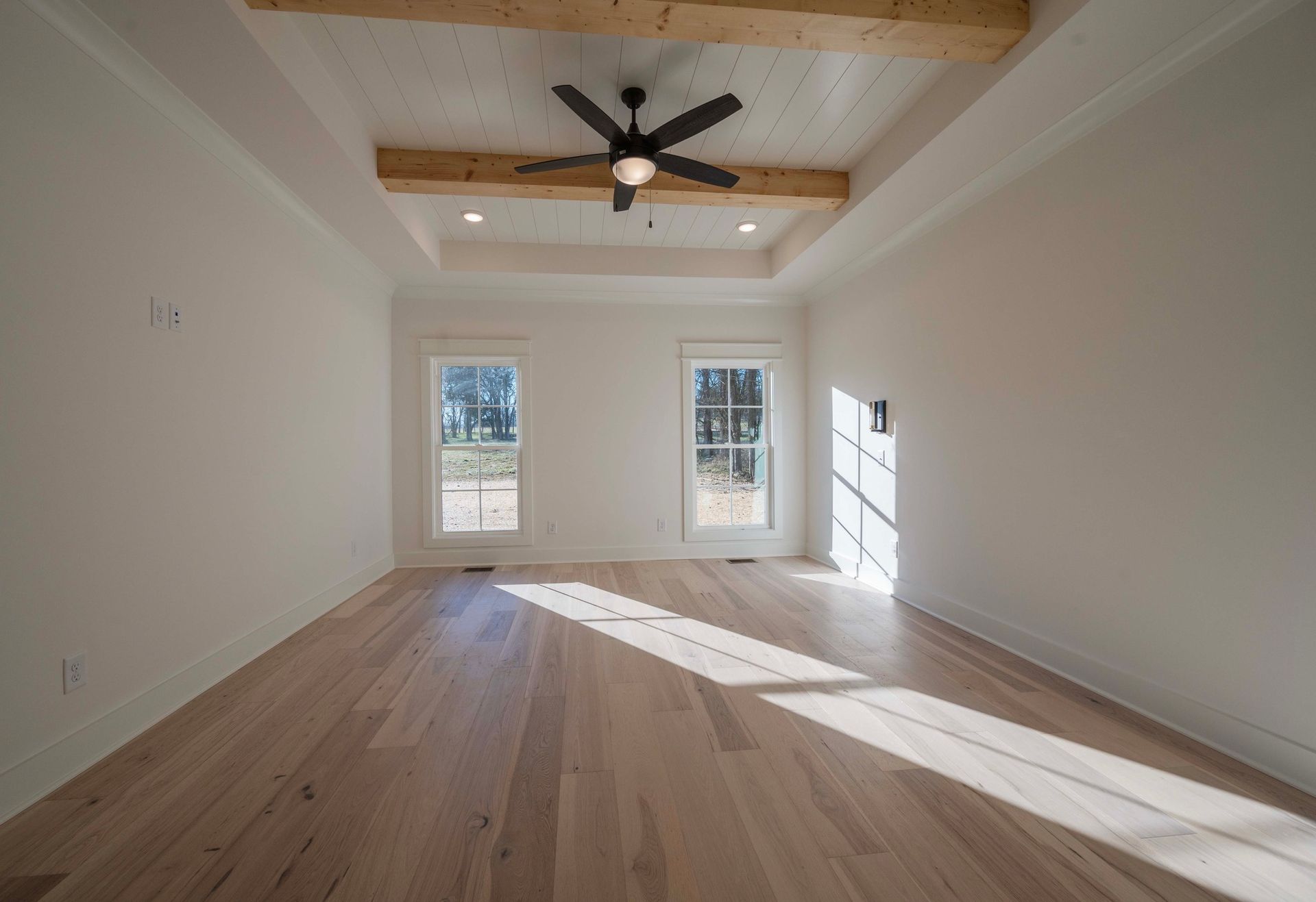 Empty room with light wood floors, white walls, and a ceiling fan. Two windows let in sunlight.