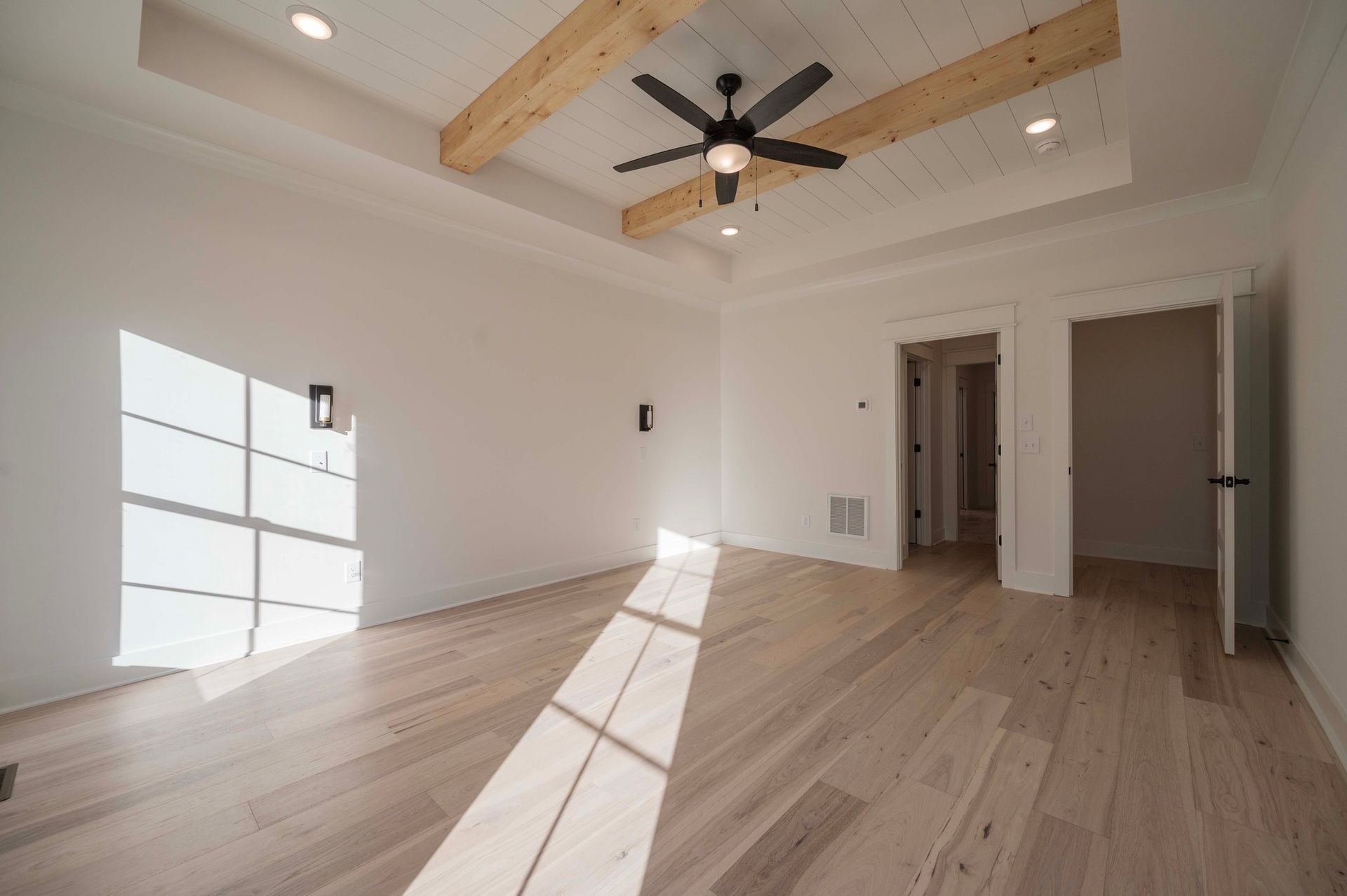 Empty bedroom with wood floor, white walls, and a ceiling fan. Natural light shines in from the left.