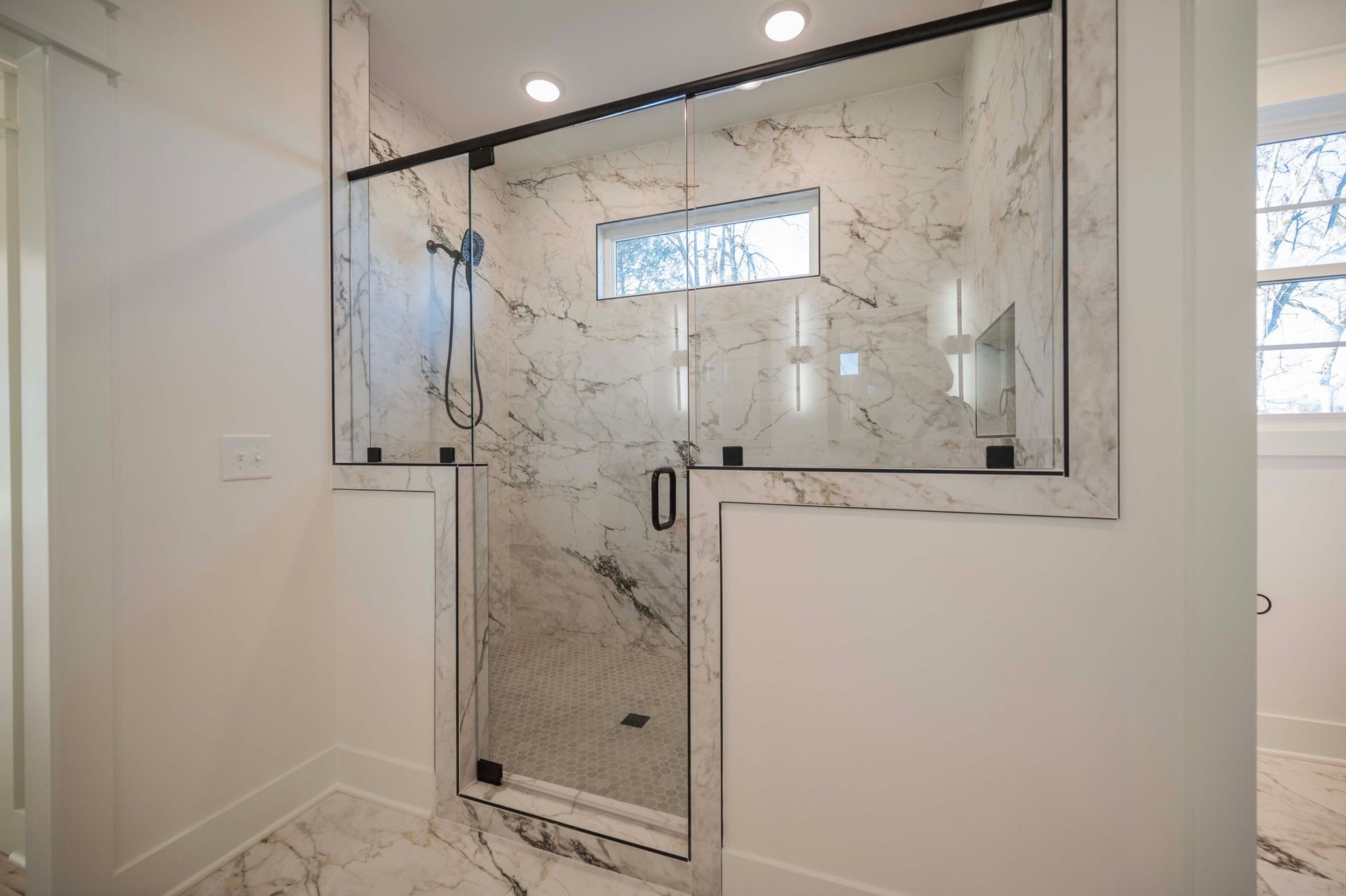Modern bathroom with marble shower and black-framed glass door, white walls, and window.
