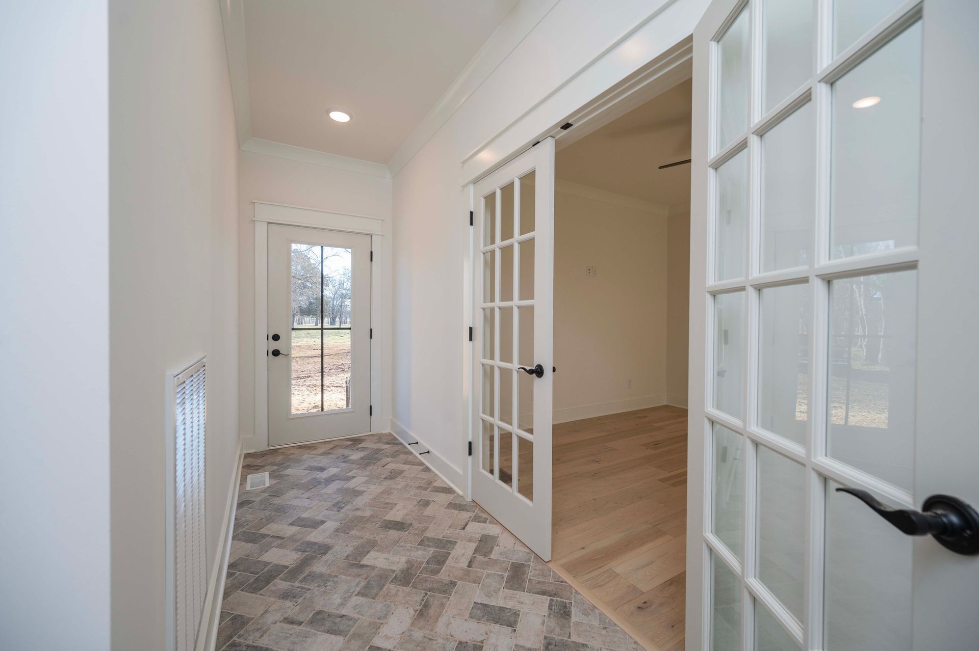 Hallway with herringbone floor, a door on the left, and French doors on the right leading to a room.