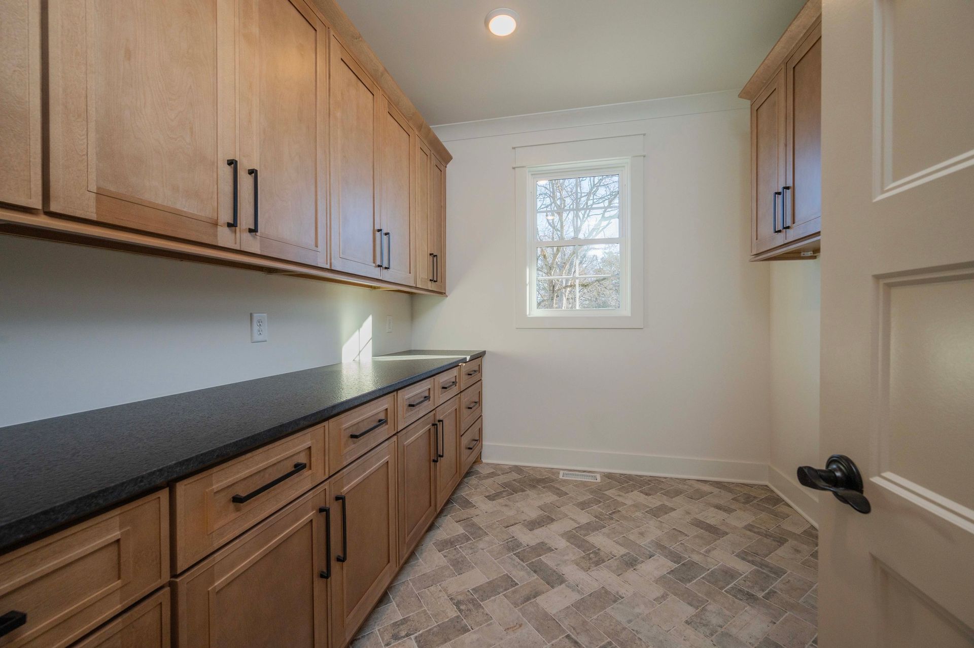 Pantry with light wood cabinets, dark countertop, and herringbone tile floor. A small window lets in light.