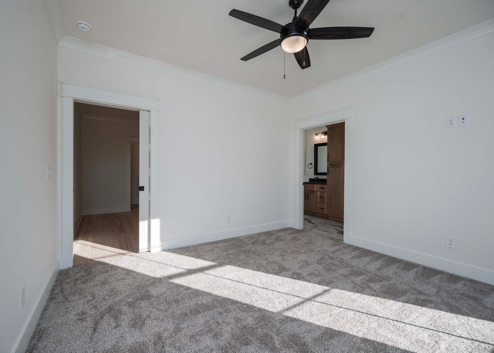 Empty bedroom with gray carpet, white walls, and two doorways. Ceiling fan. Sunlight.