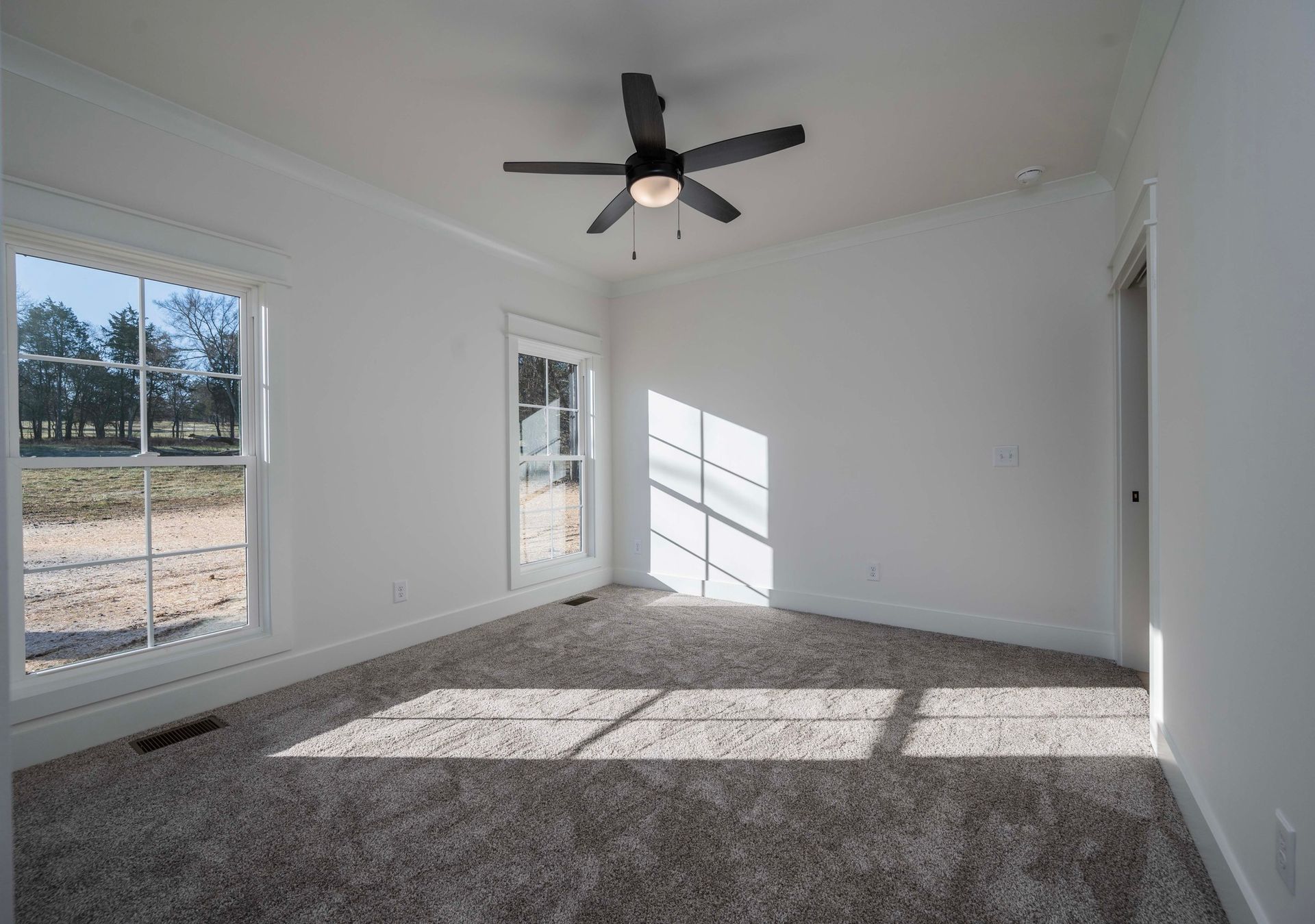 Empty room with white walls, carpet, window, and ceiling fan casting shadows.
