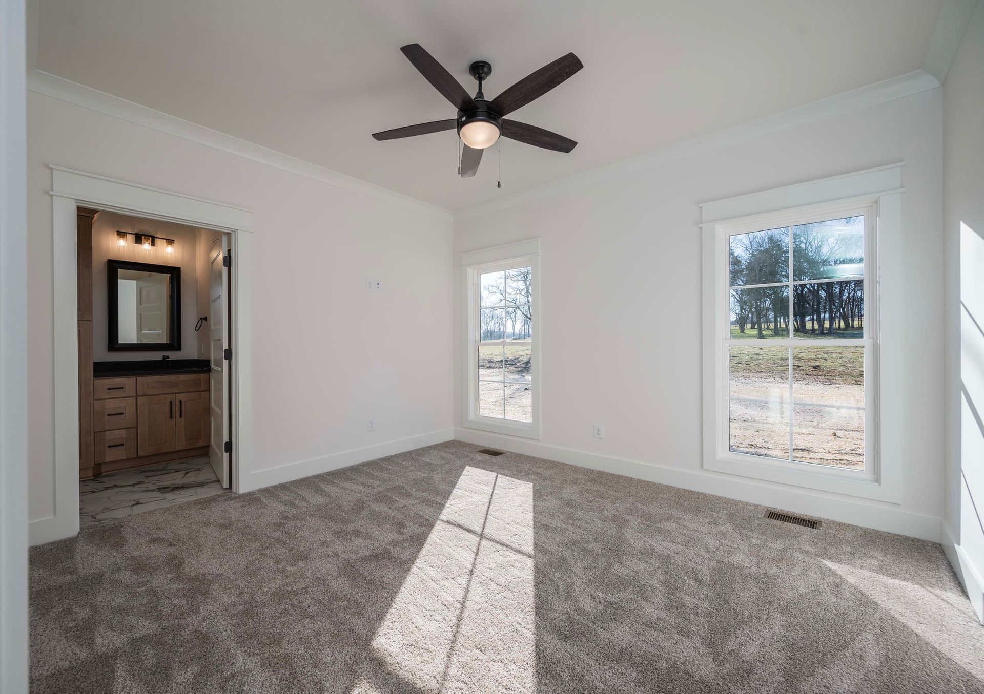 Empty bedroom with gray carpet, white walls, two windows, and bathroom entry.