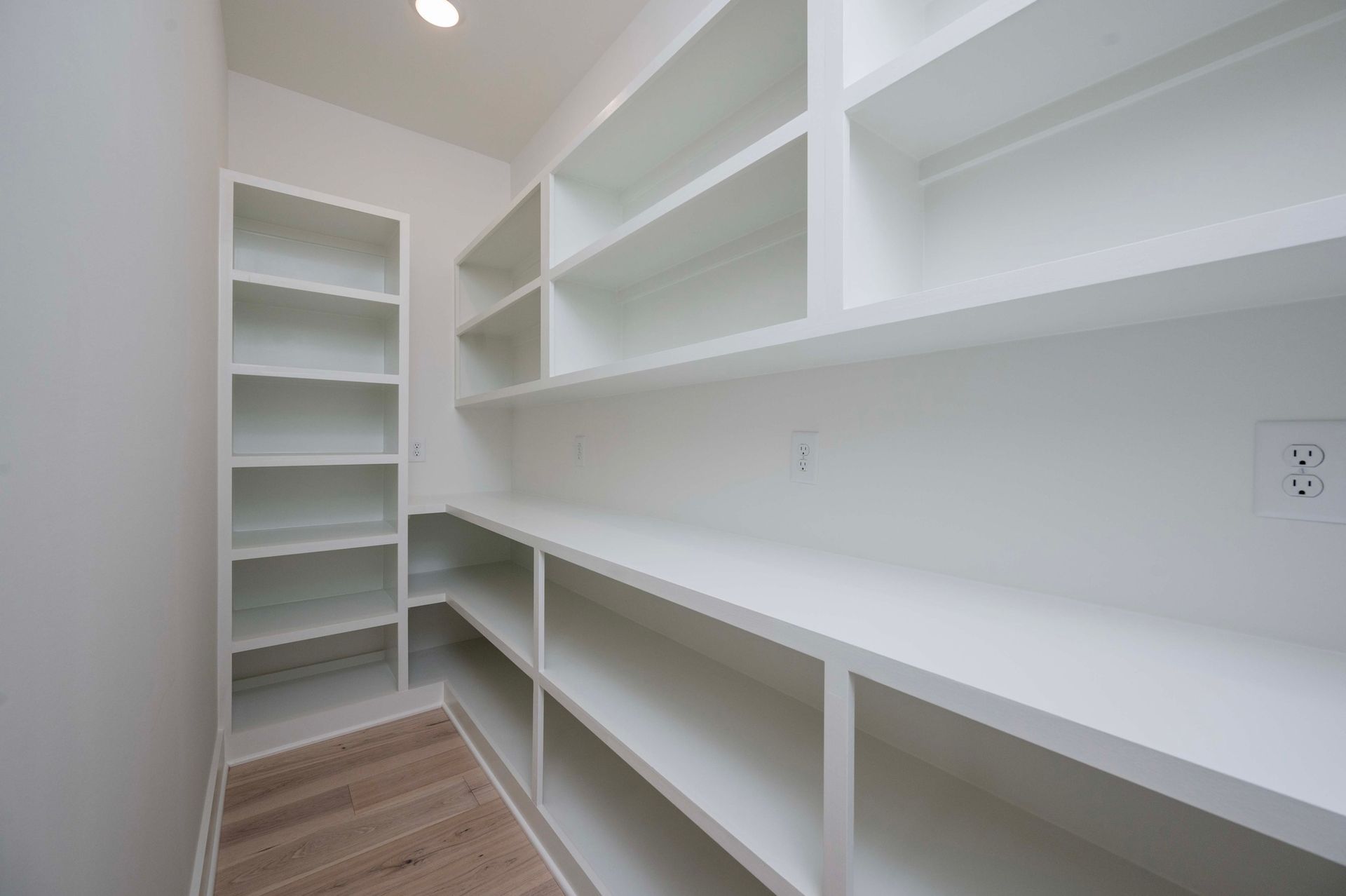 White pantry with built-in shelves of varying heights. Empty, wooden floor, bright overhead light, and an electrical outlet.