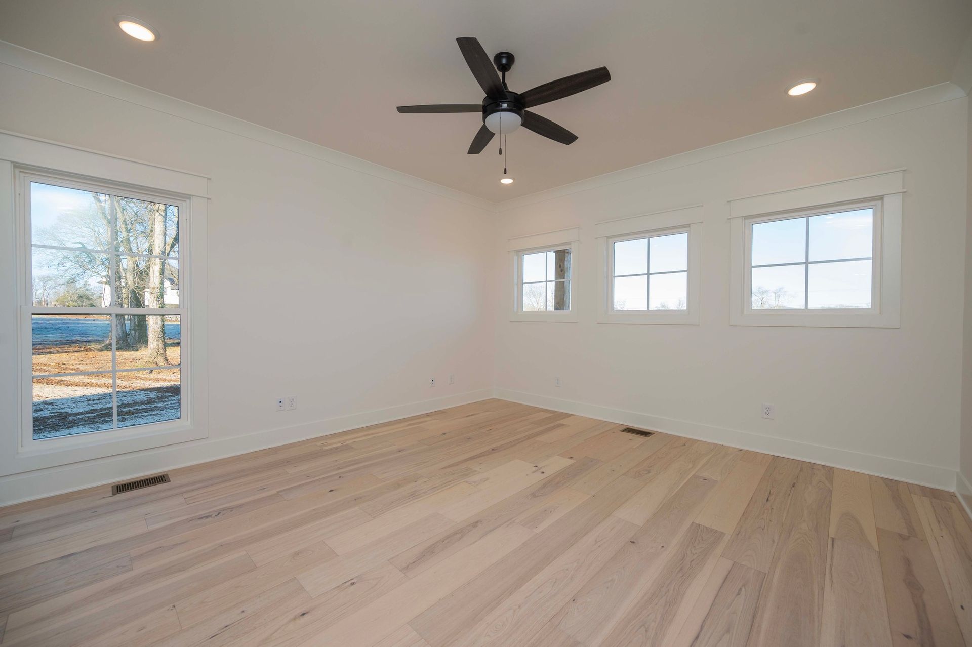 Empty room with light wood floor, white walls, and a ceiling fan. Large window and three smaller windows.