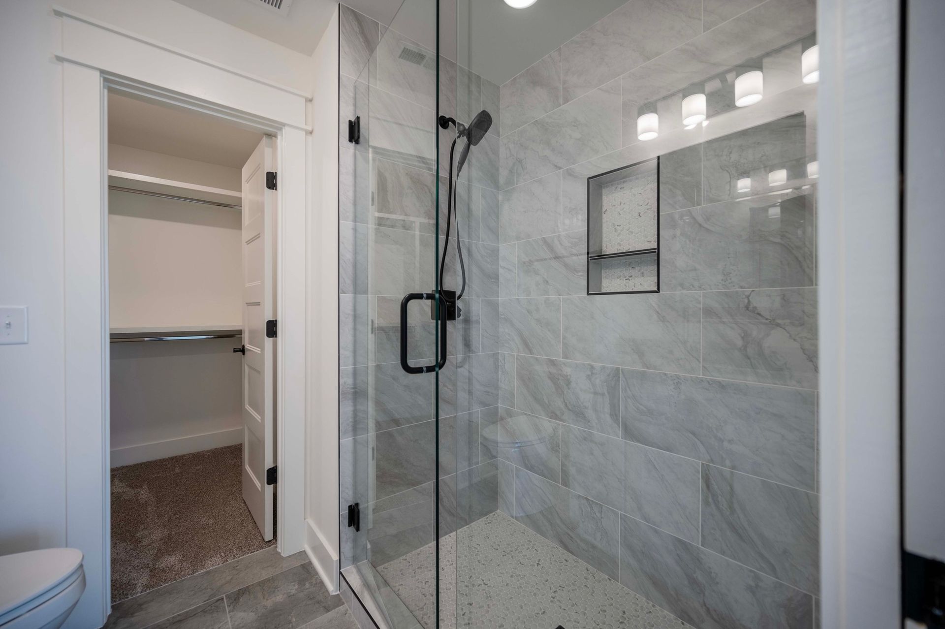 Bathroom with a glass shower, gray tiled walls, and a walk-in closet with shelves.
