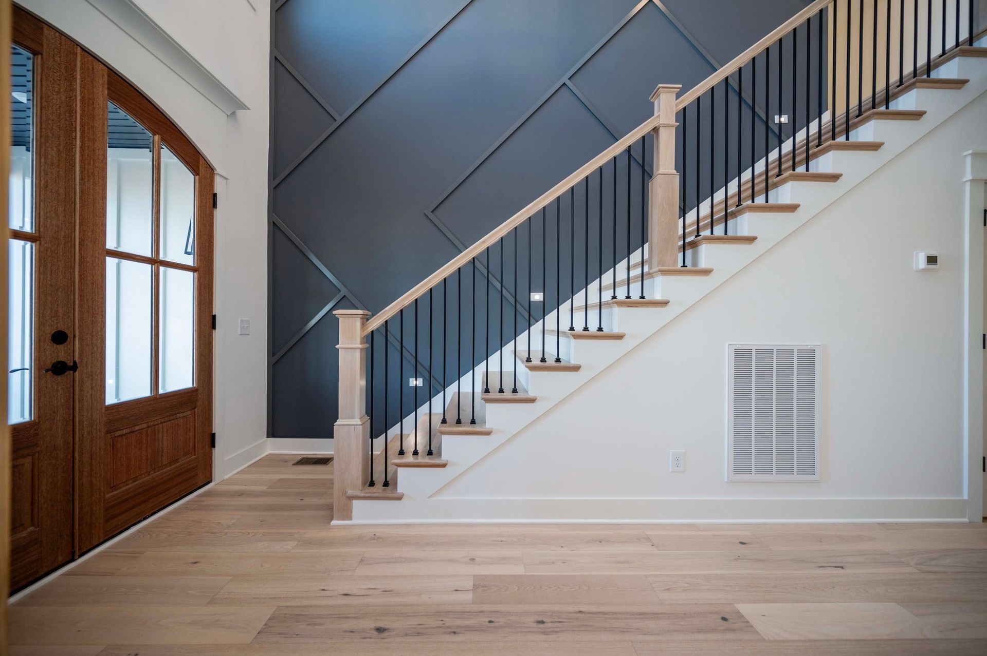 Wooden door and staircase with black railing and light wood floor against white and gray walls.