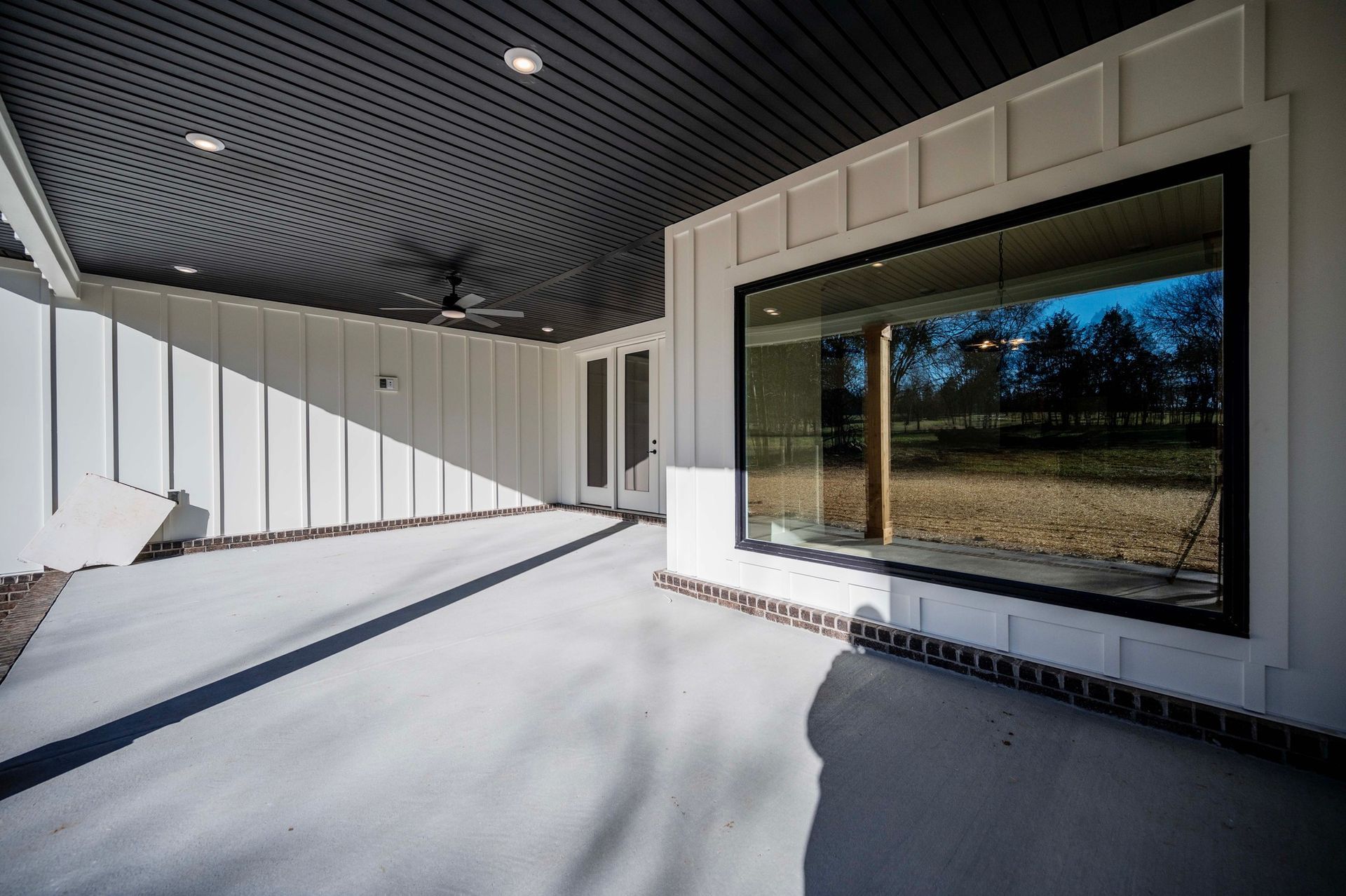 Covered patio with large window reflecting trees. White walls, grey floor, and dark ceiling.