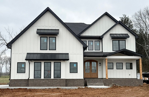 White farmhouse with black trim, awnings, and roof. Wooden door, brick base, and front porch.