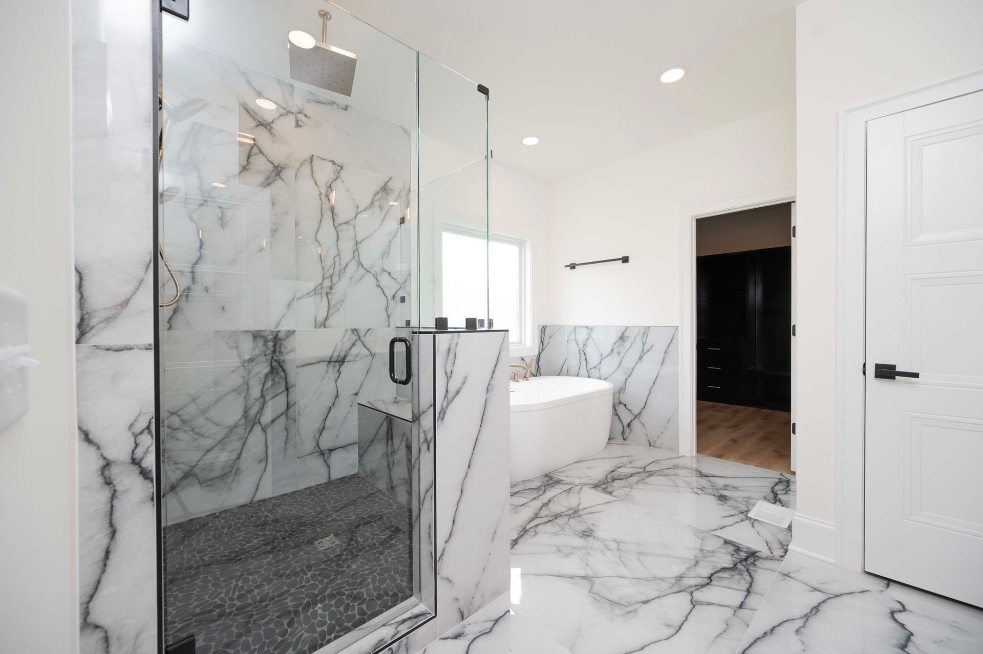 Luxurious bathroom with marble shower, flooring, and tub. White walls, dark door and fixtures.