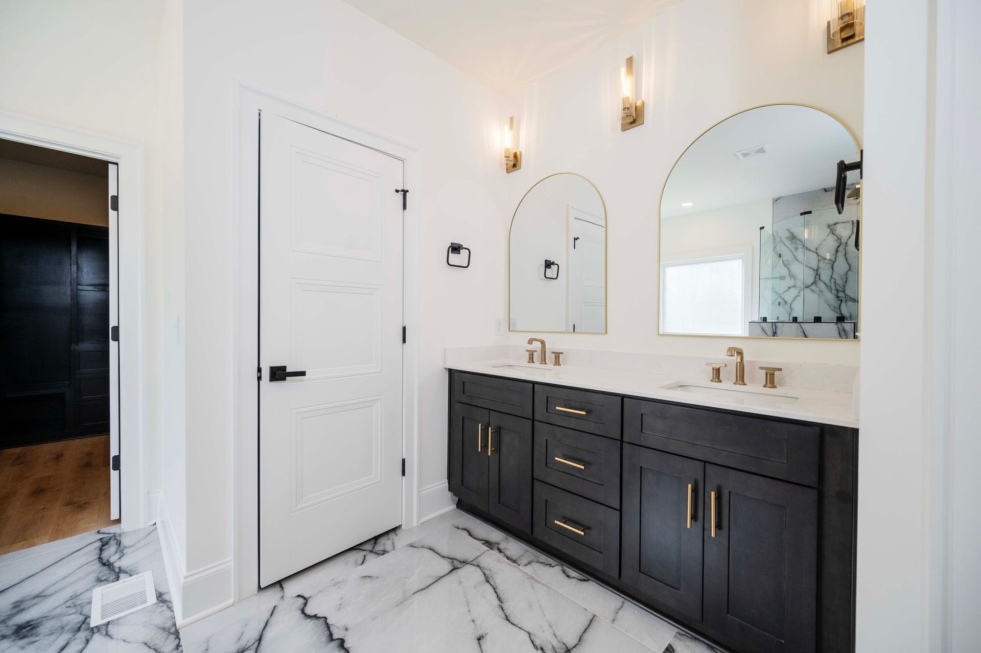 Modern bathroom with black vanity, white marble floor, arched mirrors, and gold fixtures.